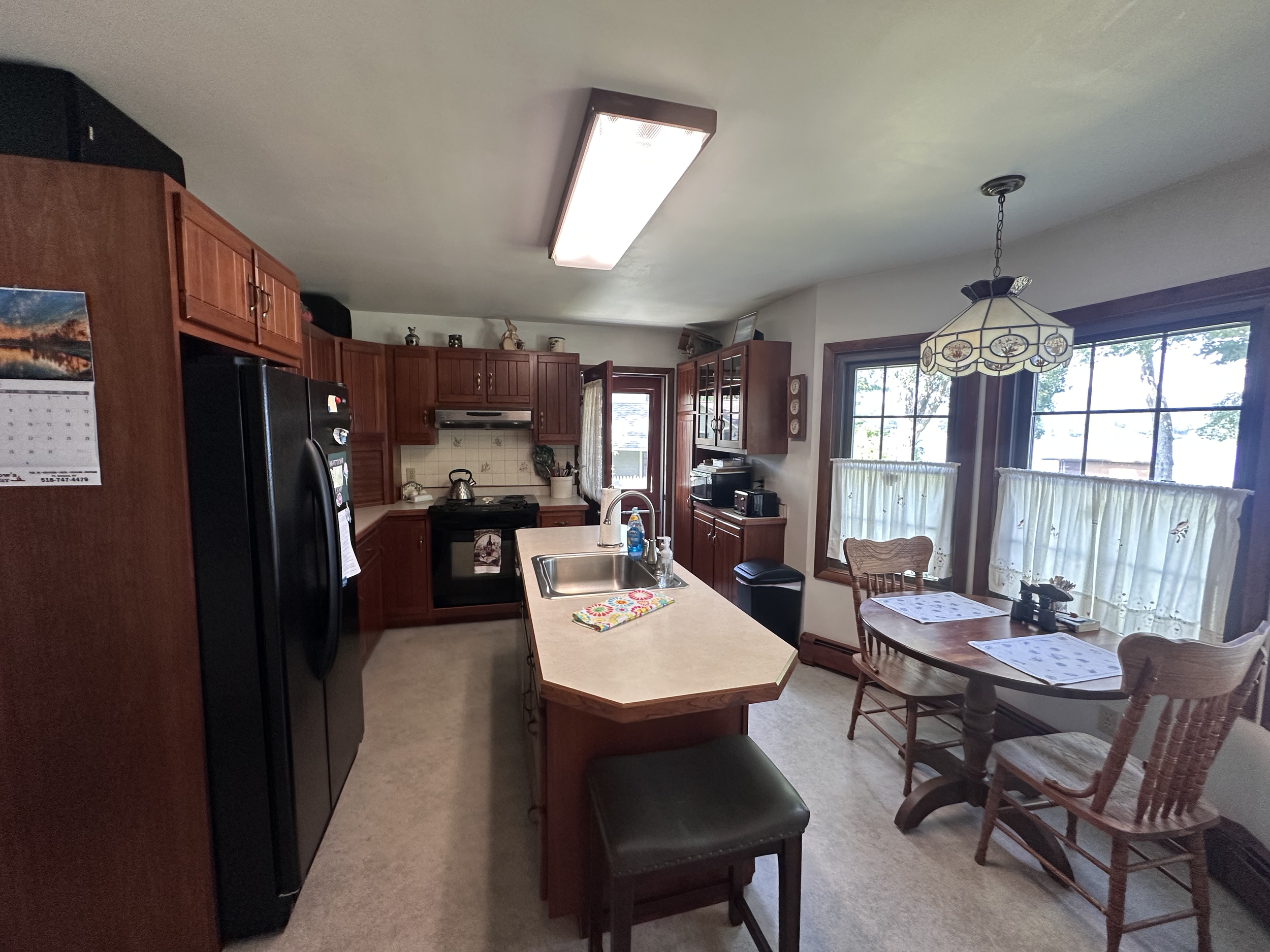 Kitchen with wooden cabinets, black refrigerator, stove, sink on island, and dining table by windows with lace curtains and a stained glass hanging light