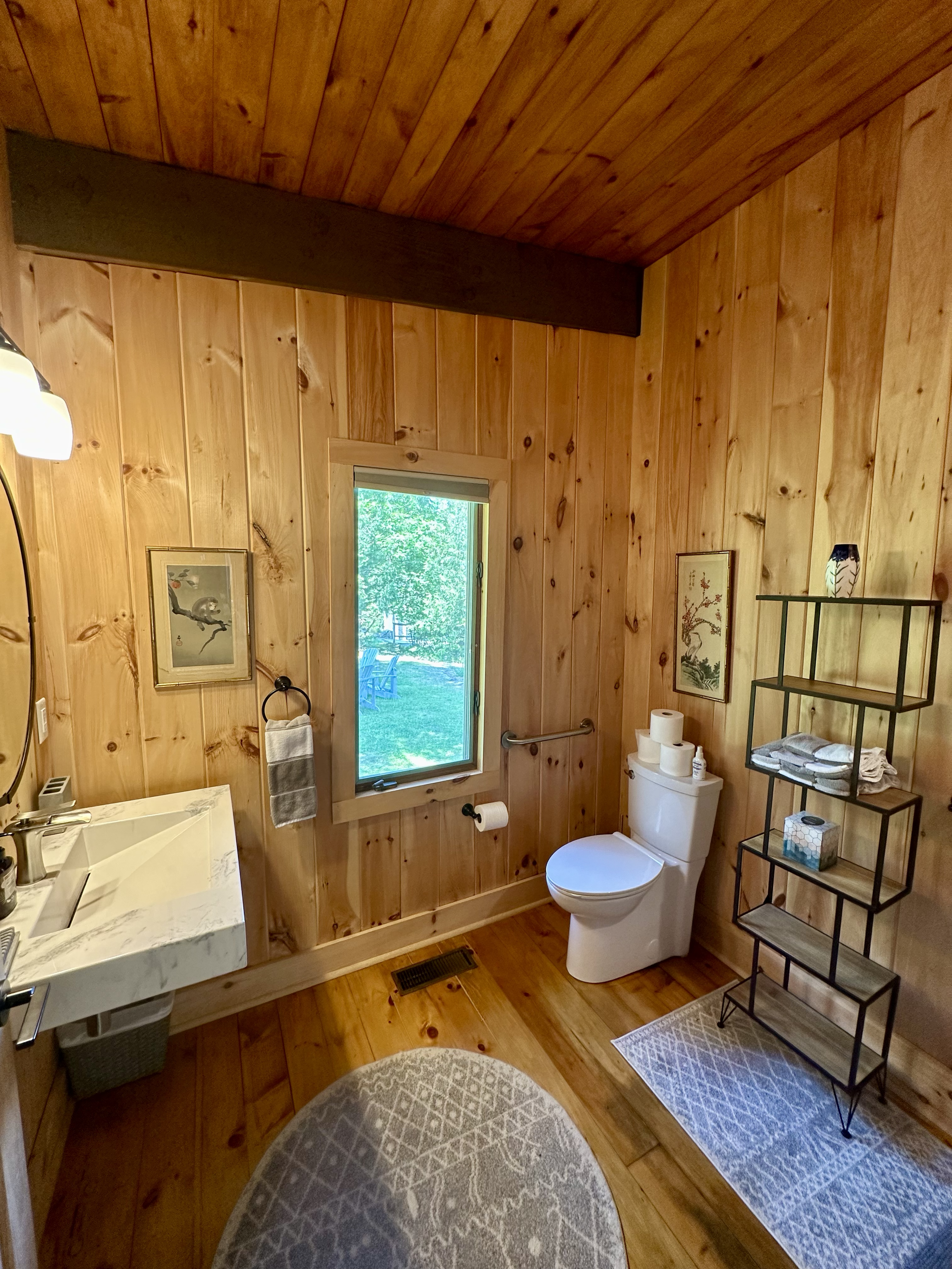A rustic bathroom with wood paneling walls and ceiling, a white toilet, a small window with a view of greenery outside, a marble sink, and a black metal shelving unit with towels, tissues, and decorative items.