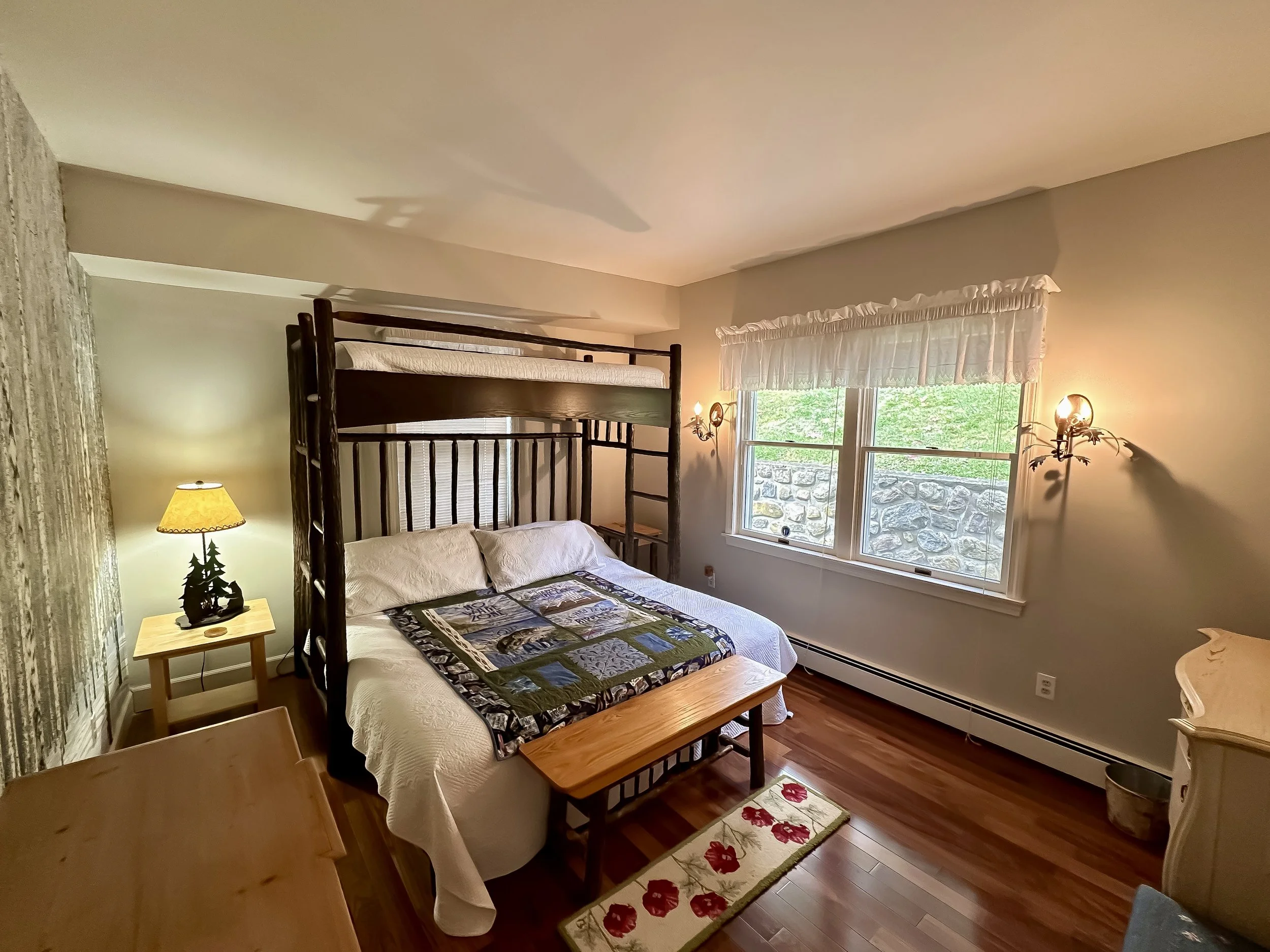 A cozy bedroom with a double bed, a wooden bench at the foot, a bunk bed frame above, a window with a ruffled valance, a small side table with a lamp and decorative trees, and light fixtures on the wall.
