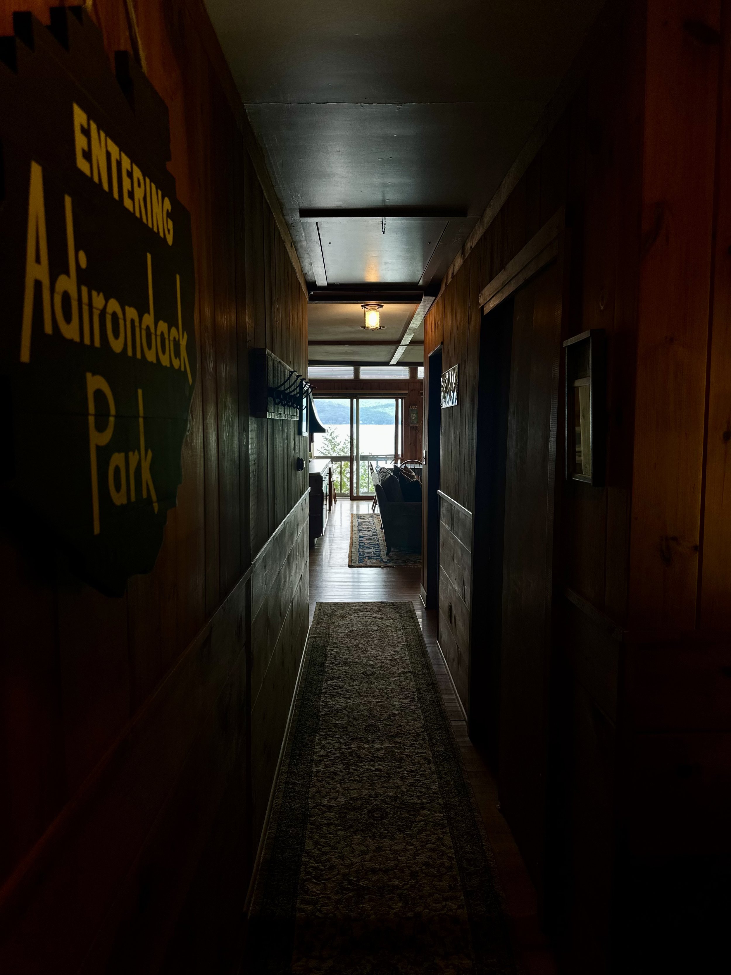 A narrow indoor hallway with wooden paneled walls leading to a bright living area with a view of a lake outside.