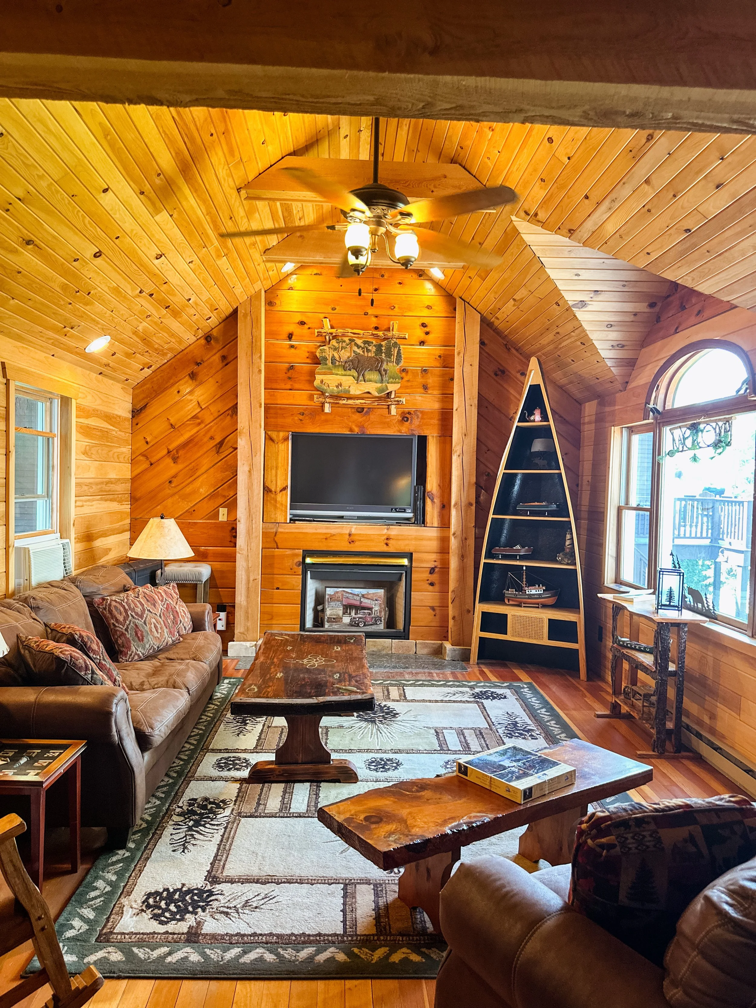 Living room with wood-paneled walls and vaulted ceiling, brown leather sofa with patterned pillows, wooden coffee table, area rug with tree and nature motifs, fireplace, mounted TV, ceiling fan, large windows letting in natural light, and nautical de