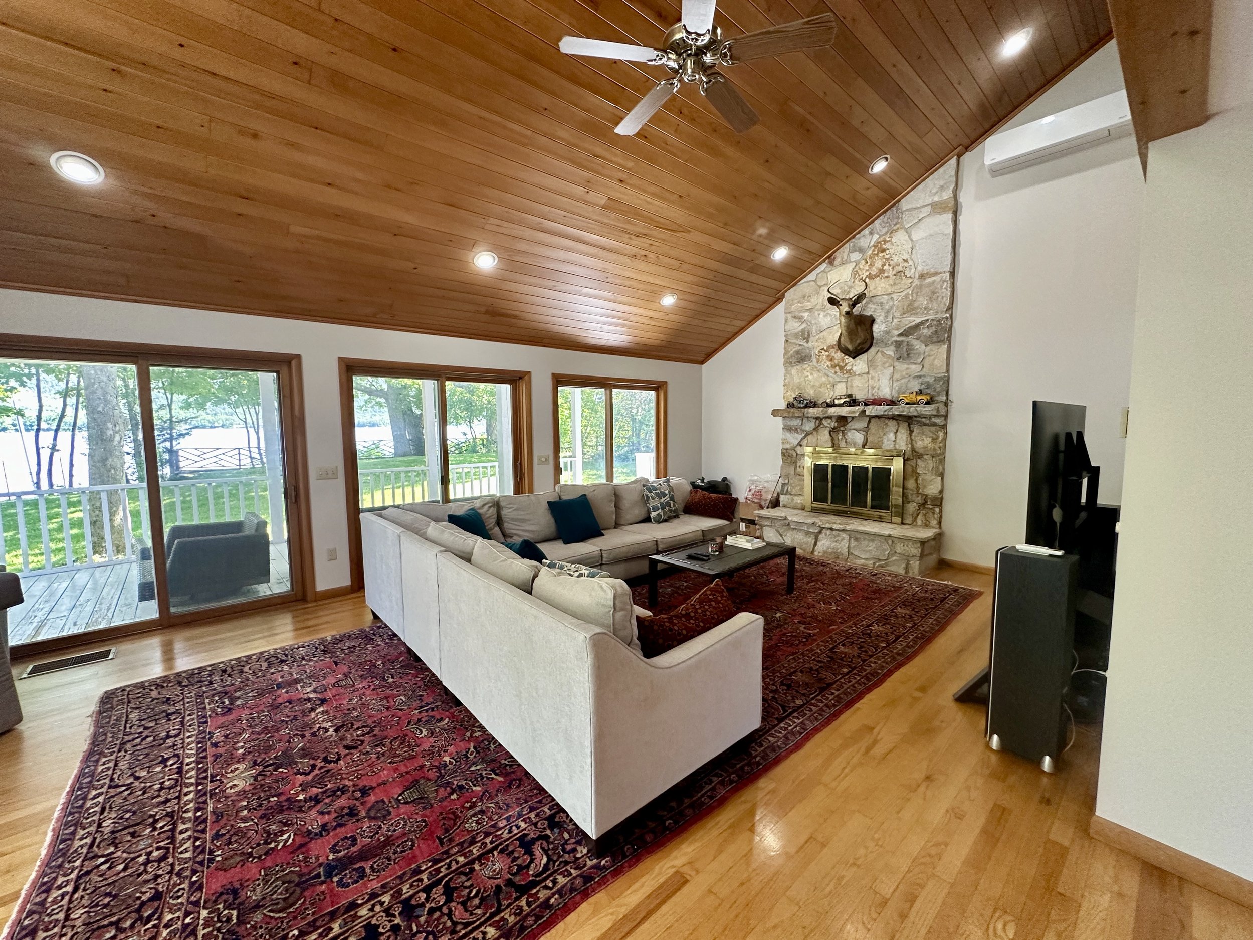 Living room with a vaulted wooden ceiling, a stone fireplace with a mounted deer head, a sectional sofa, a patterned area rug, glass sliding doors leading outside, and a ceiling fan.