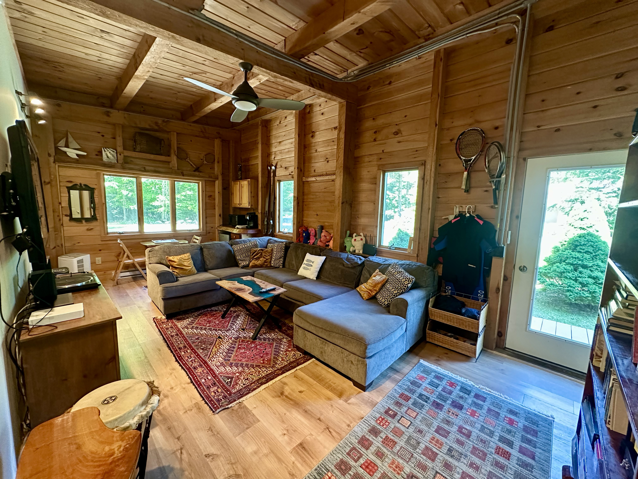Living room with wooden walls and ceiling, gray sectional sofa with yellow and patterned throw pillows, wooden coffee table, large window with green trees outside, ceiling fan, and various decor items.