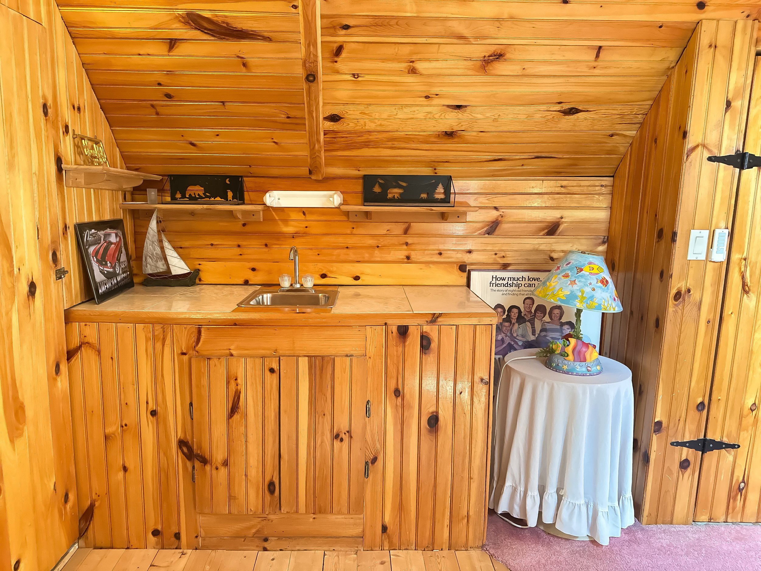 Interior of a wood-paneled room with a kitchen counter, small sink, and various decorations including framed pictures, a model sailboat, and a lamp with a colorful patterned shade.