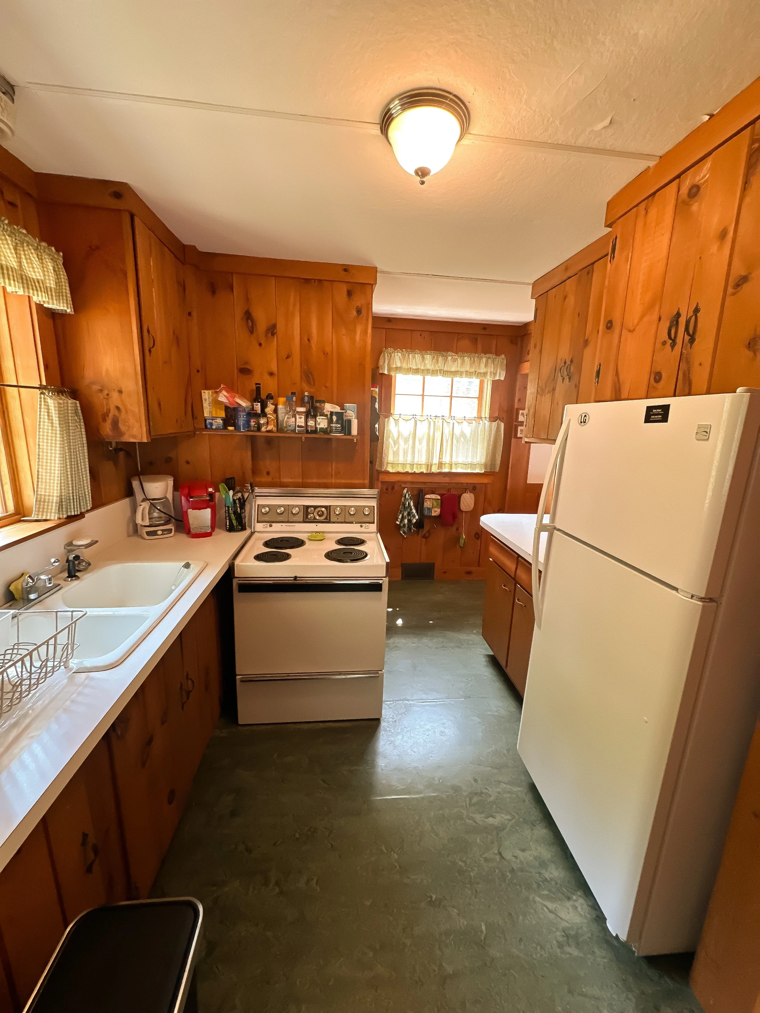 Small kitchen with wooden cabinets, white refrigerator, stove, and sink. Windows with yellow curtains, countertop appliances, and wall-mounted bottle shelf.