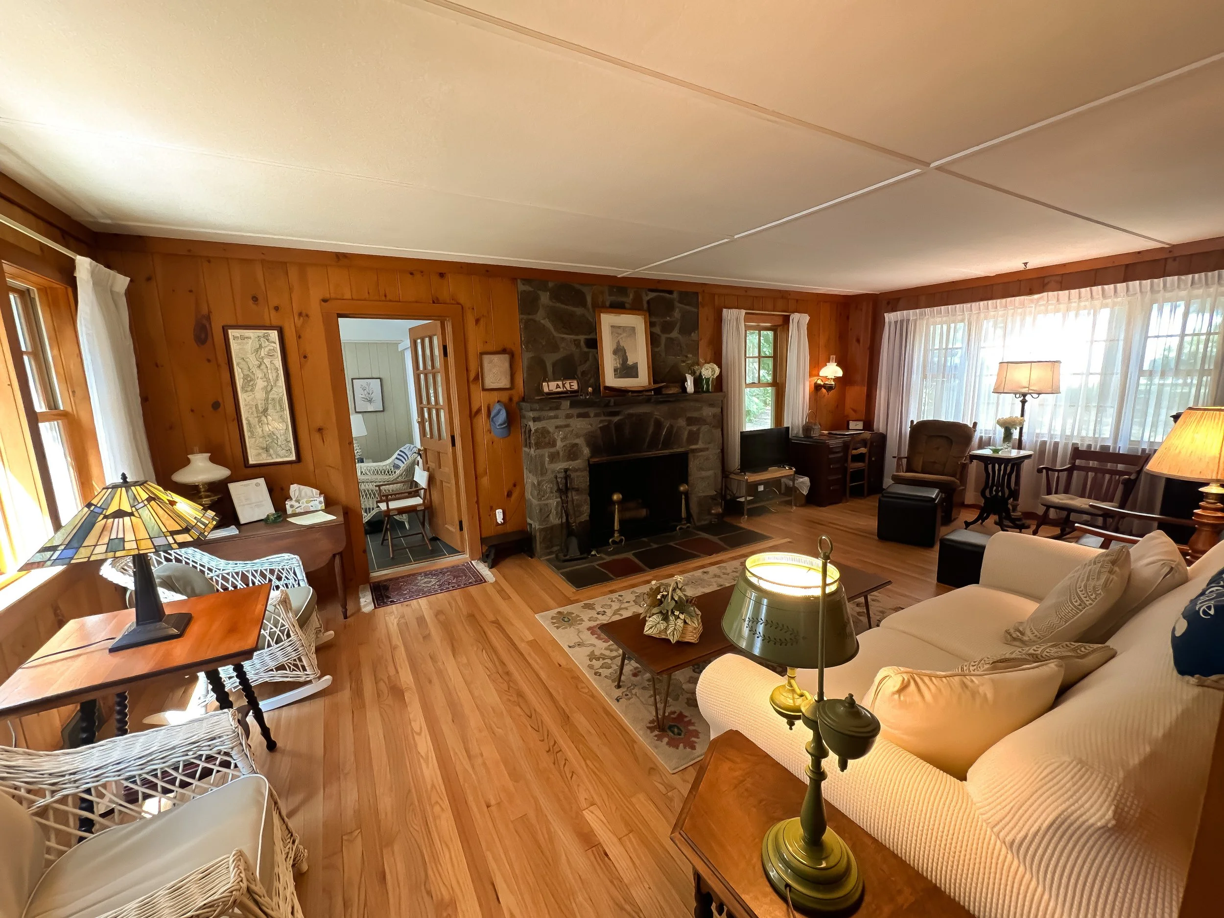 A cozy living room with wood-paneled walls, a stone fireplace, and large windows with white curtains. The room has a mix of furniture including a white sofa, wicker chairs, and various side tables with lamps. The hardwood floor is partially covered b