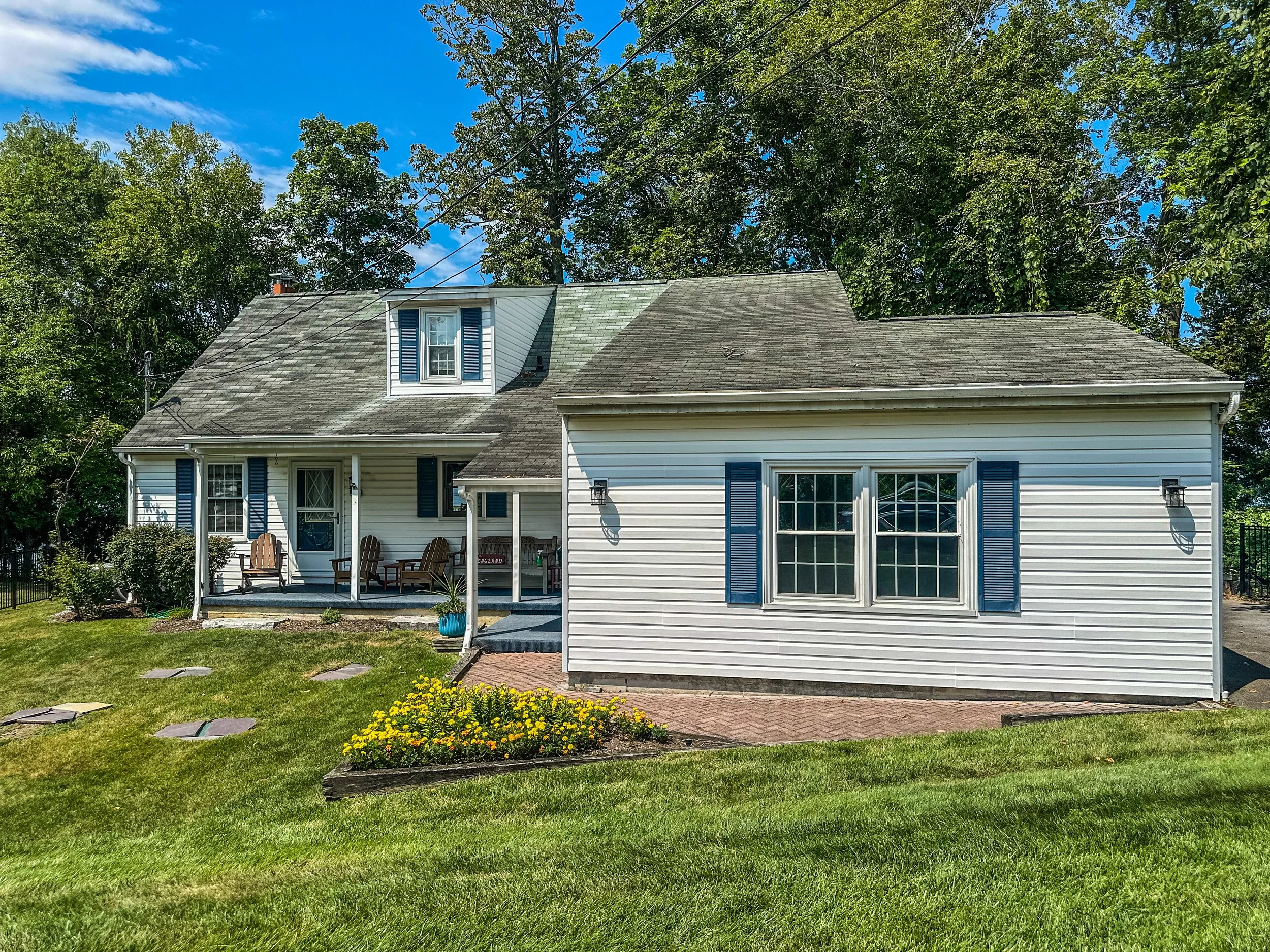 A white house with blue shutters and a porch with Adirondack chairs, surrounded by a green lawn and trees, on a sunny day.