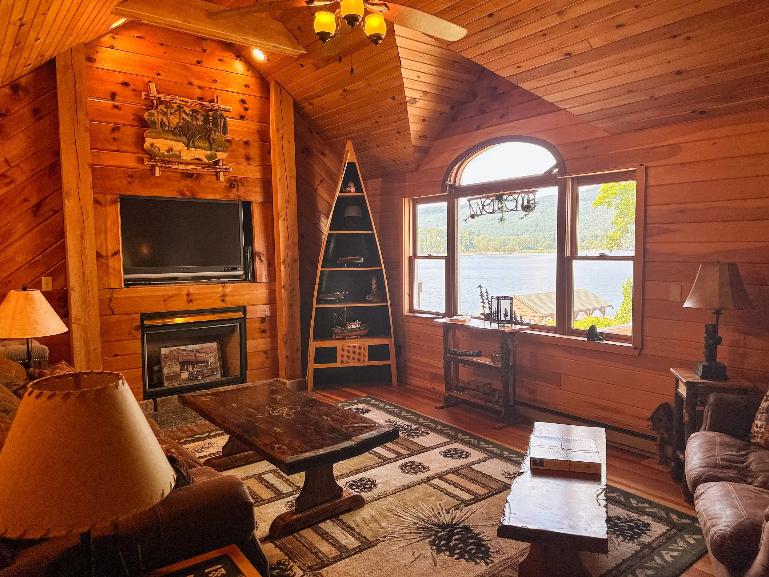 A cozy wooden living room with a large window revealing a lake and trees outside. The room features wood-paneled walls and ceiling, with a mounted flat-screen TV above a fireplace on the left. There is a triangular black bookshelf beside the window, 