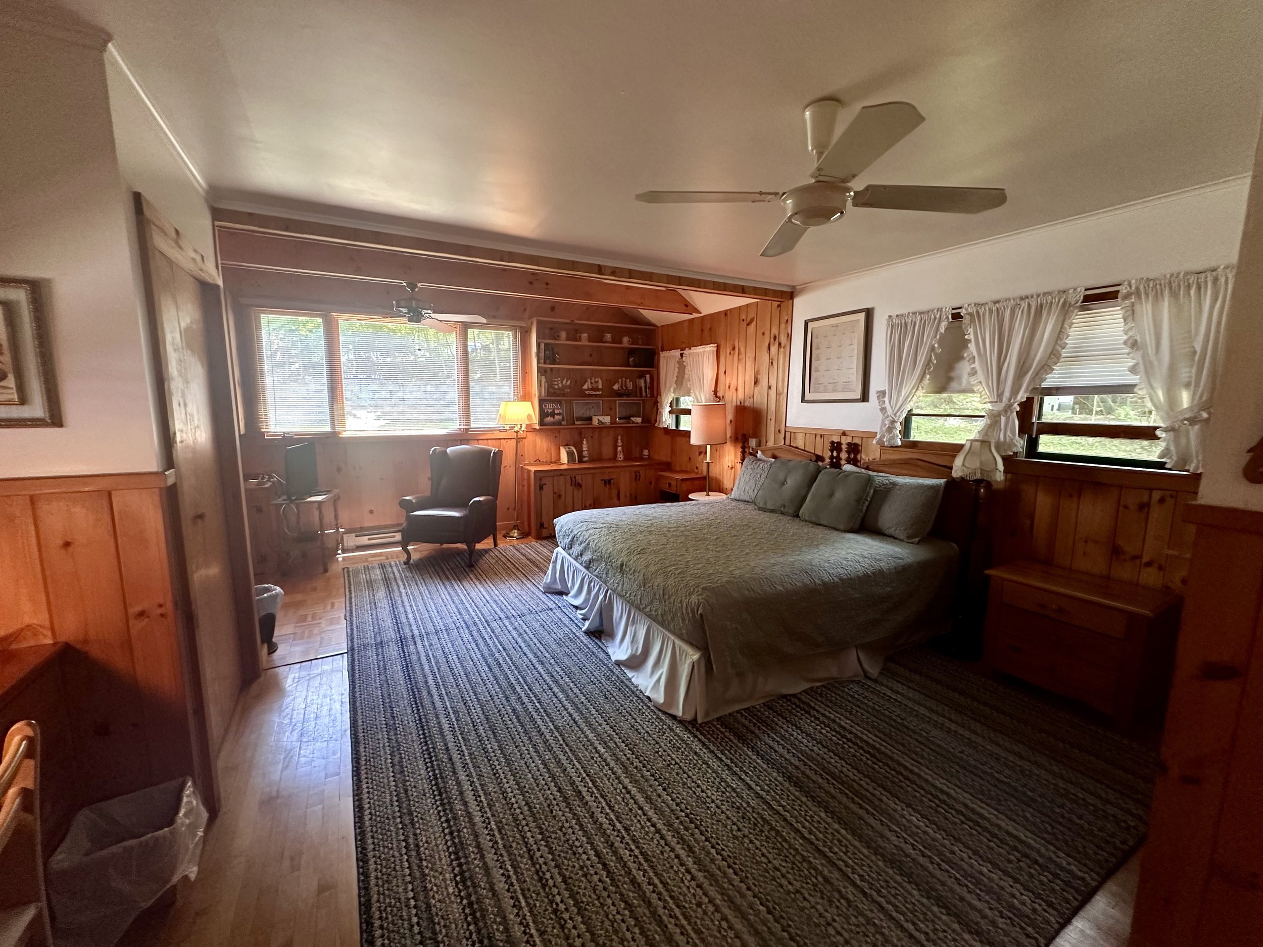 Cozy bedroom with wooden paneling, large bed with pillows, windows with white curtains, armchair, desk, bookshelf, and ceiling fan.