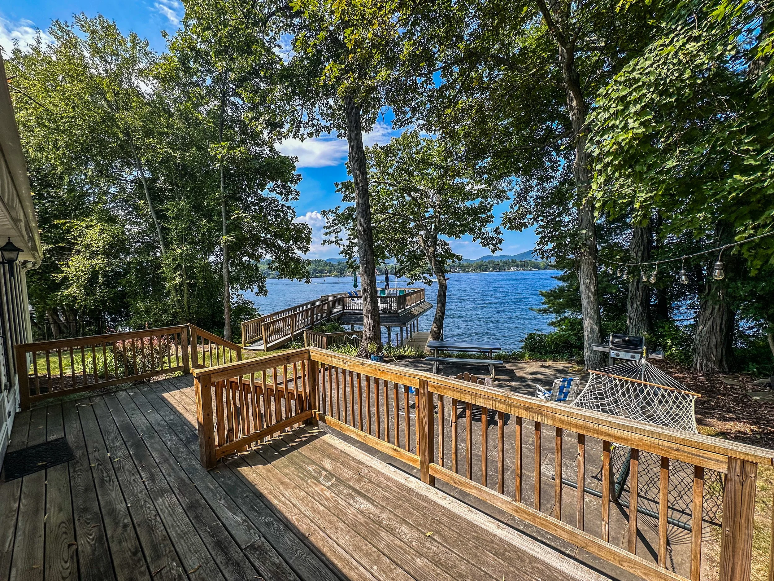 View of a lakeside backyard with wooden deck, trees, picnic tables, hammock, and a boat dock extending over the water, with mountains and a blue sky in the background.
