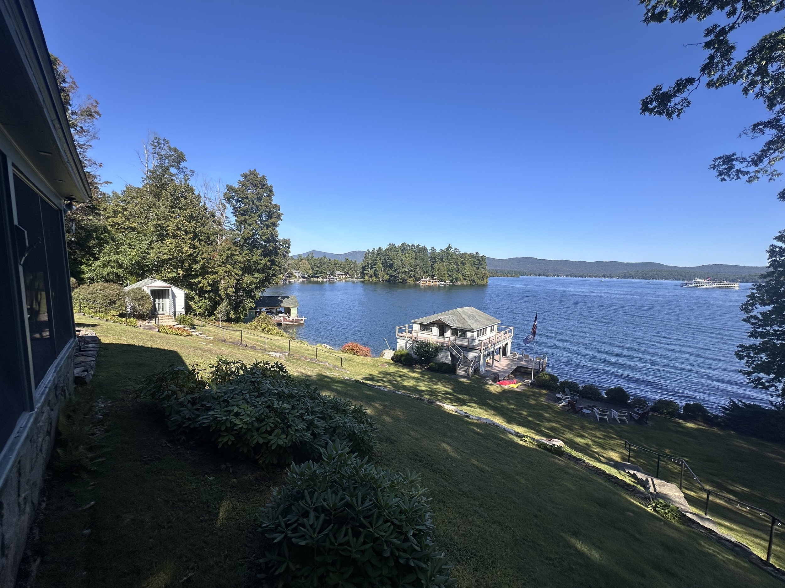 View of a lakeside property with a house on the left, a grassy lawn sloping down to the lake, a dock with a boathouse, and a boat on the water, surrounded by trees and mountains under a clear blue sky.