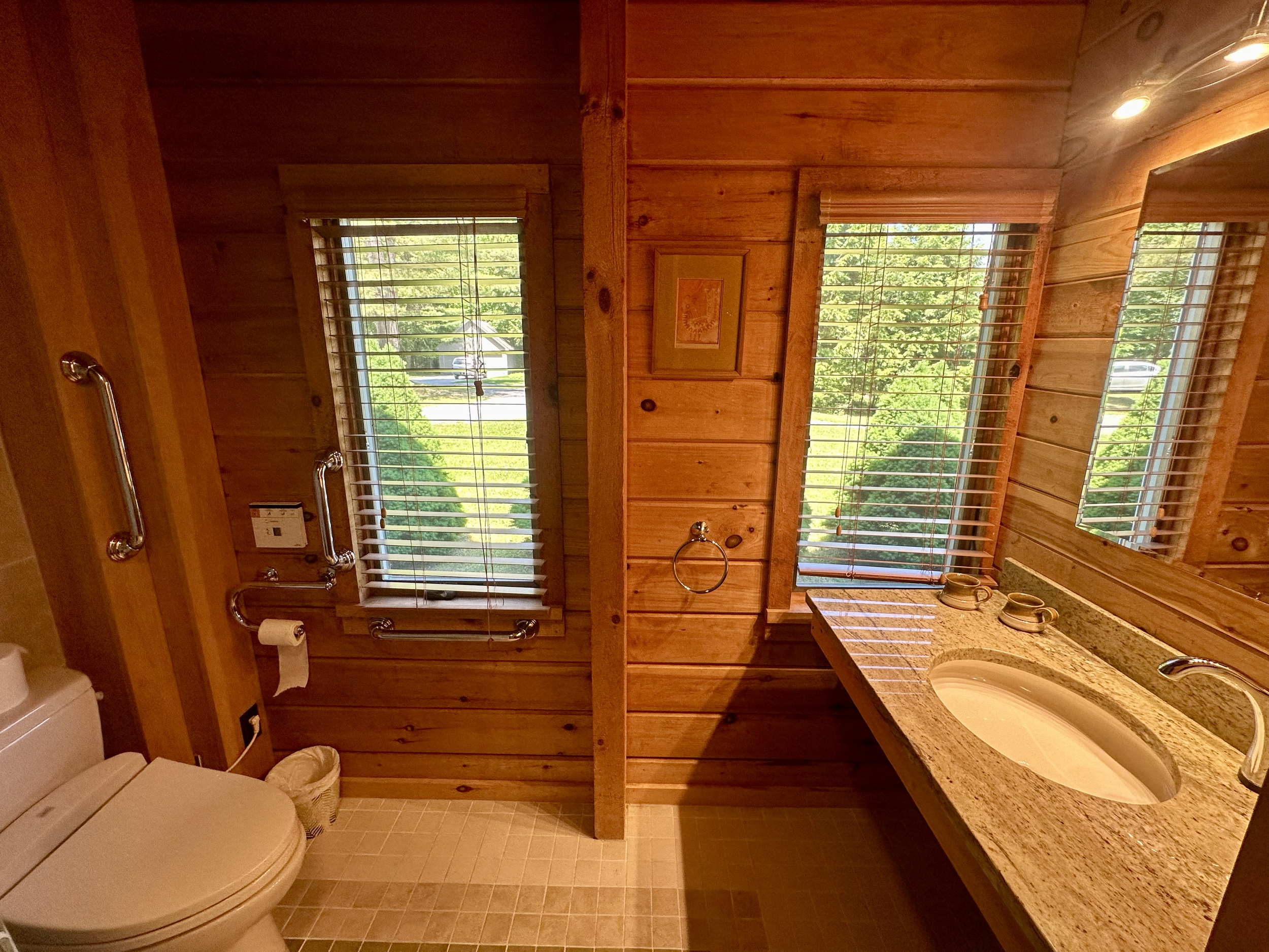 A bathroom with wooden paneled walls, a granite countertop with an oval sink, two ceramic cups, a toilet, a window with blinds, and a mirror.