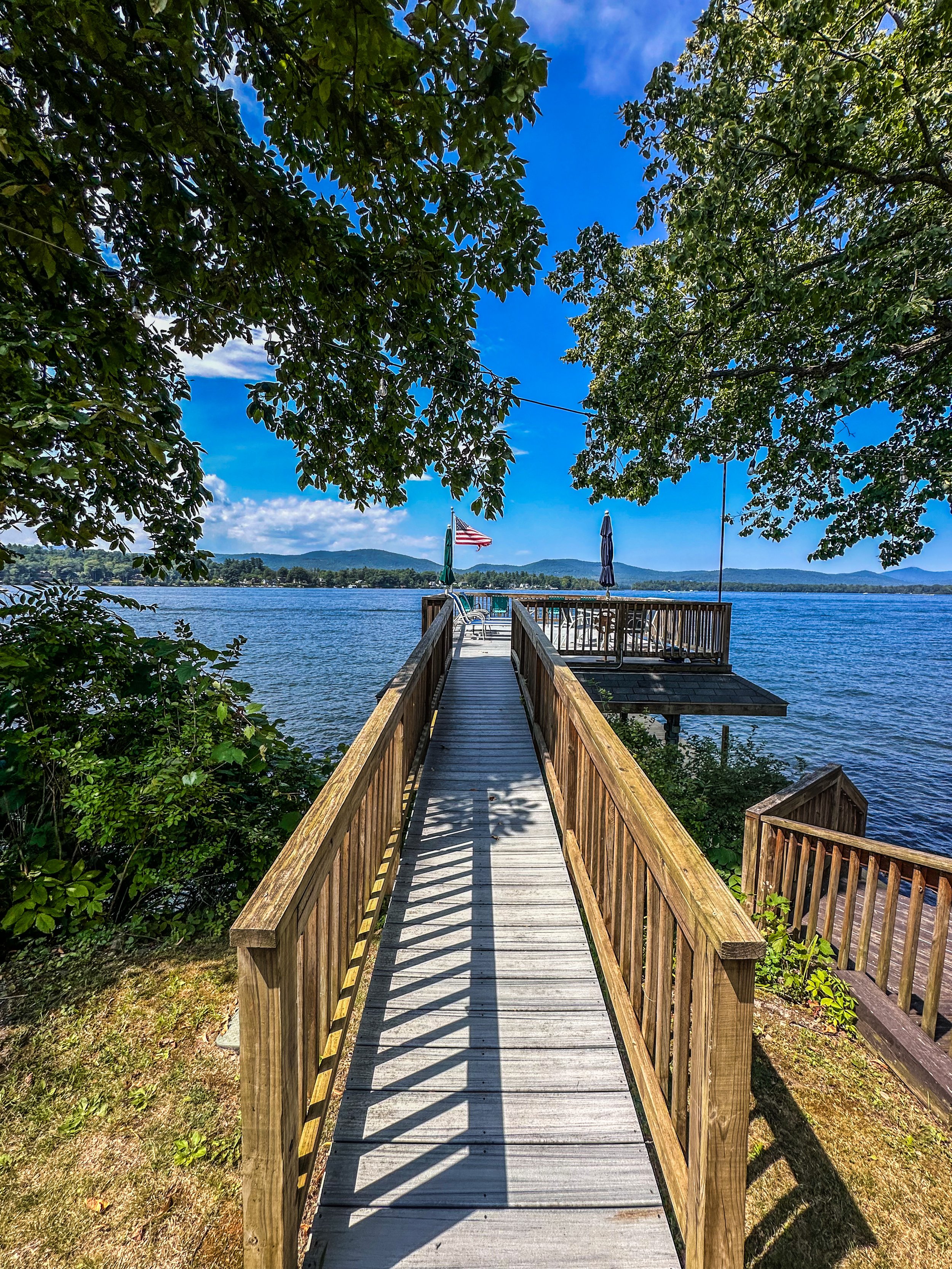 A wooden dock extending over a lake, with a small deck with umbrellas at the end, surrounded by green trees and blue sky.