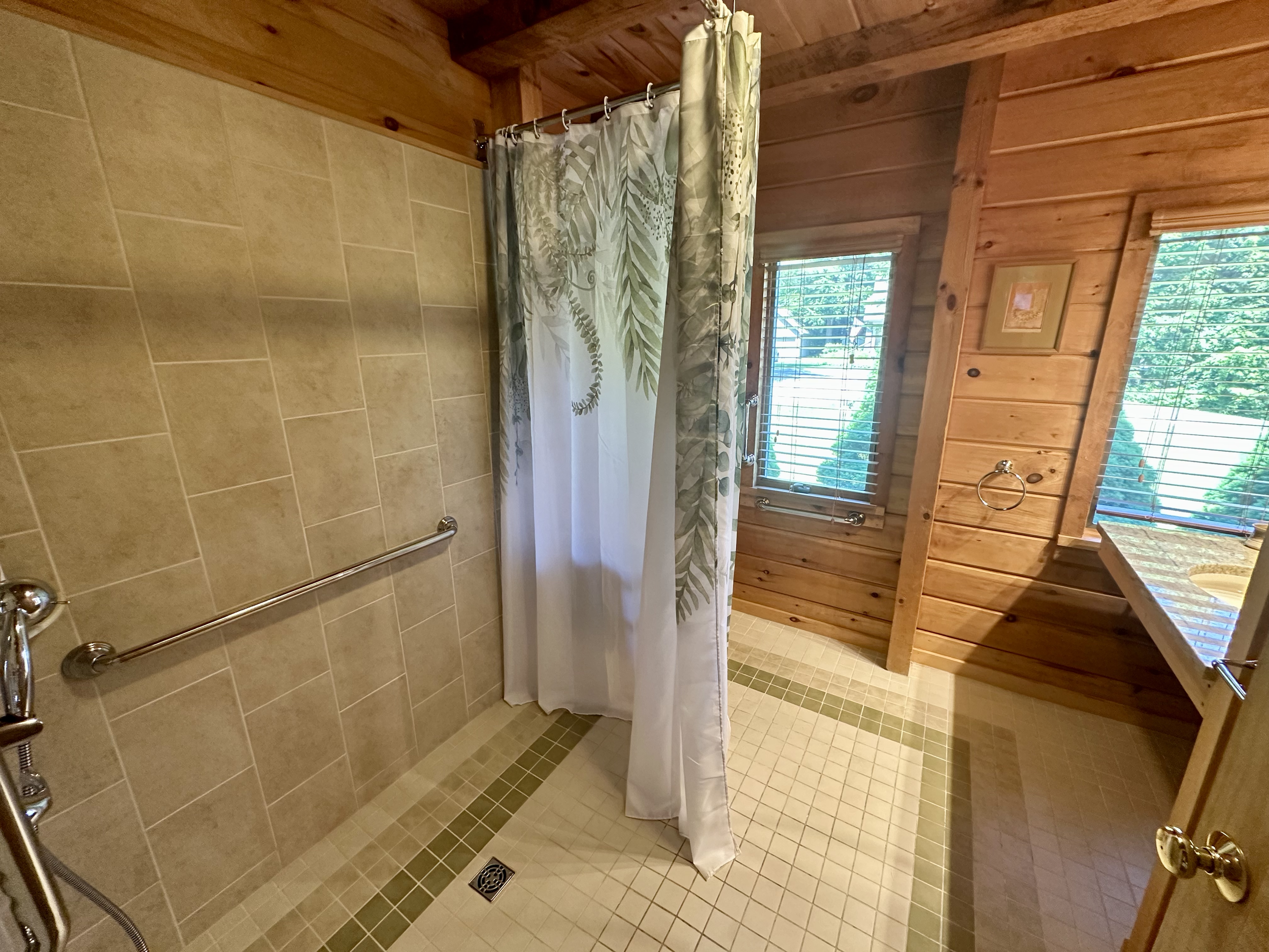 Wood-paneled bathroom with a walk-in shower, beige tiled walls, and two windows with blinds, partially covered by a curtain with a leaf pattern.