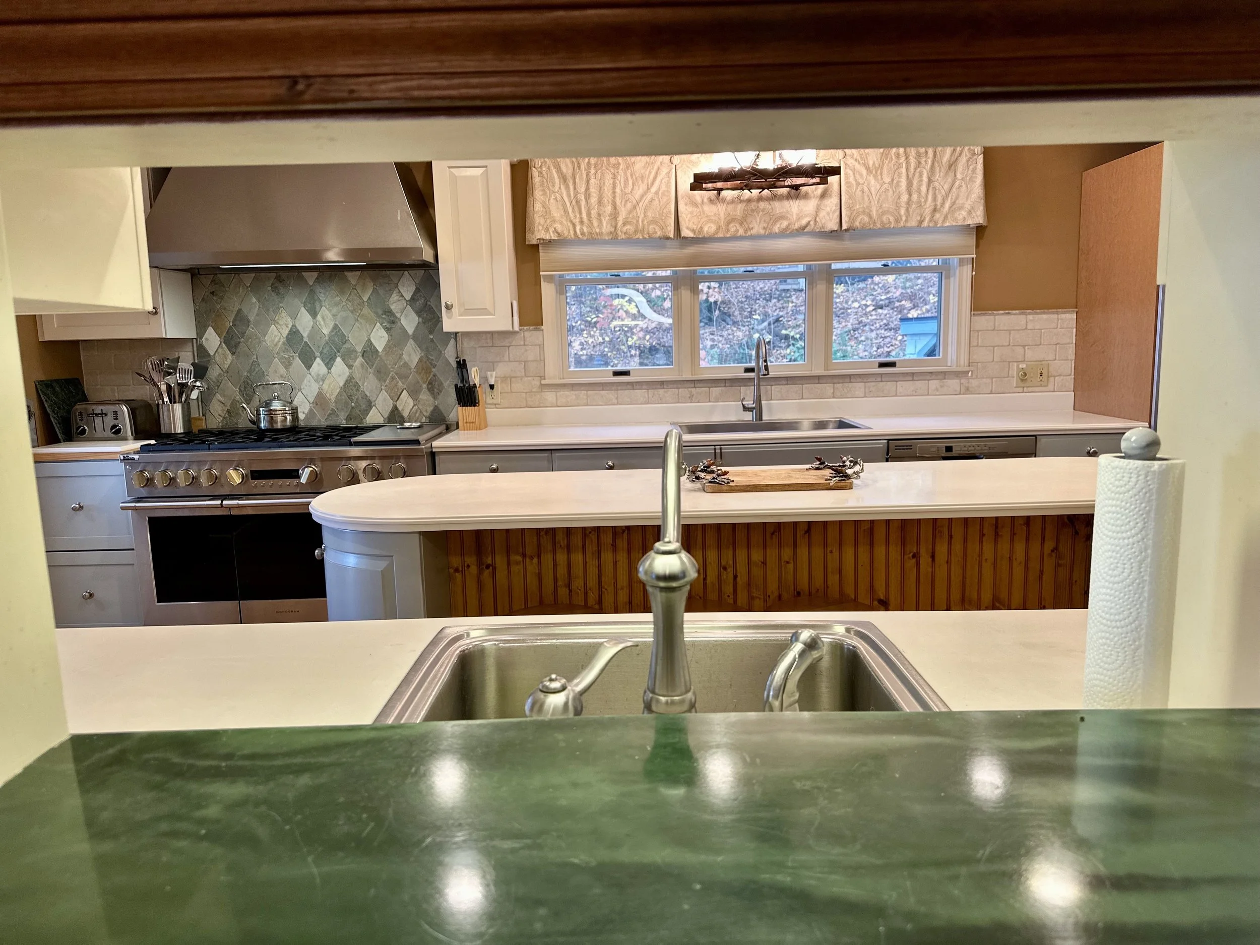 View of a kitchen through a pass-through window, showing a double sink, a tiled backsplash, a stove, and a window with a beige valance.