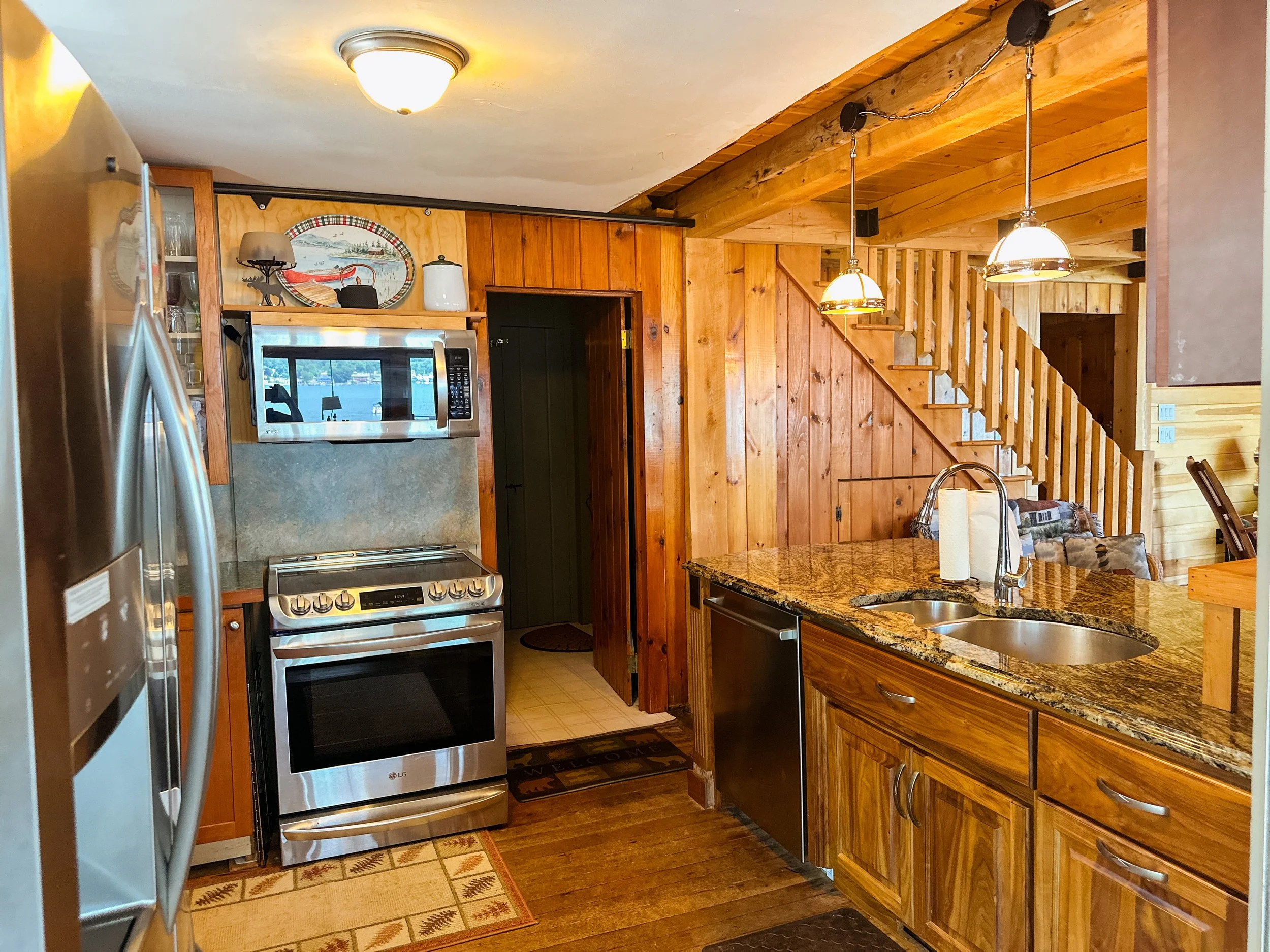 A cozy kitchen with wooden cabinets and a granite countertop, stainless steel appliances, and ceiling lights. There are stairs and a staircase railing made of wood visible in the background.
