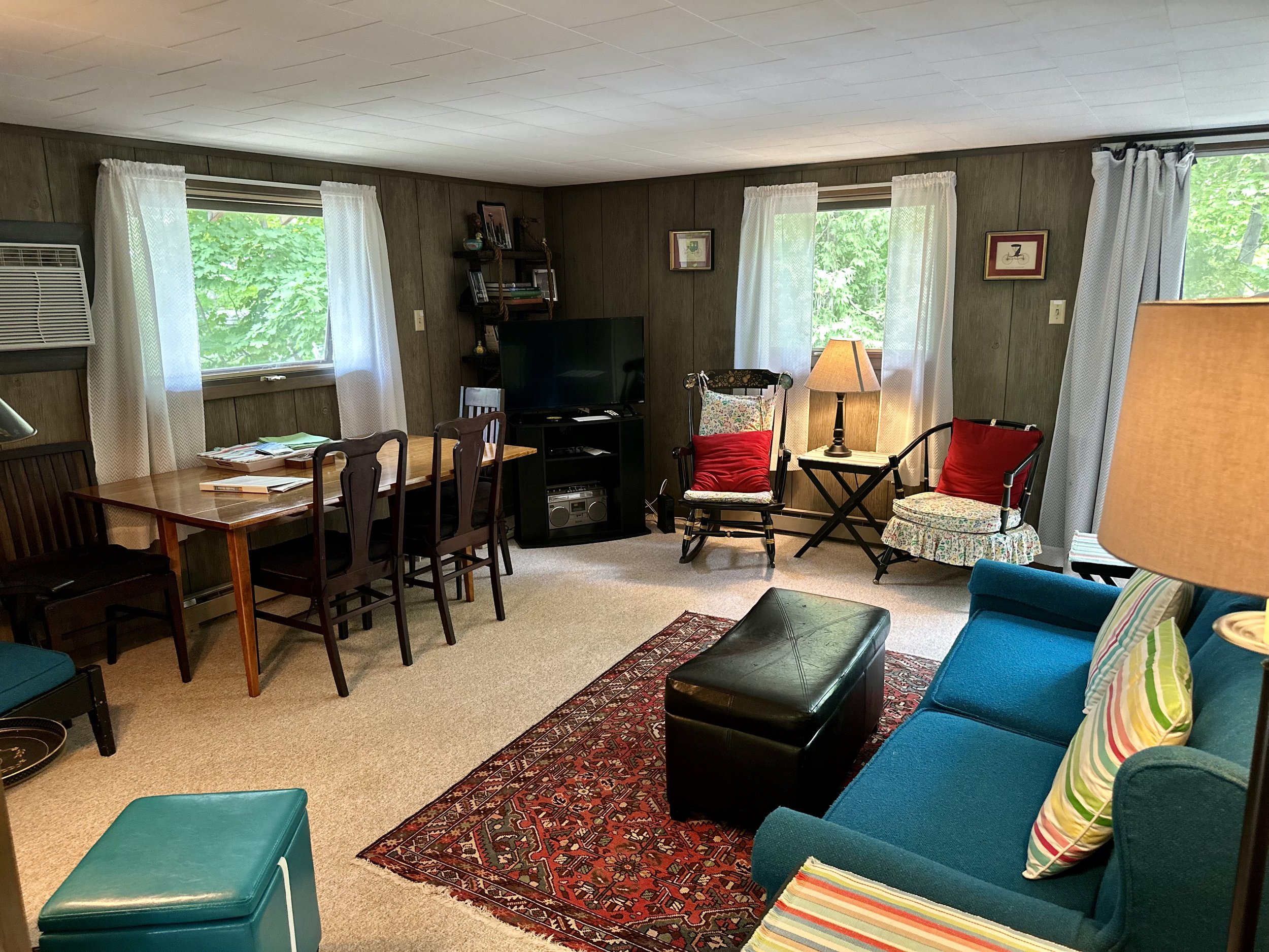 Living room with wooden walls, three windows with white curtains, blue sofa with striped pillows, black ottoman, patterned rug, armchairs with red cushions, a wooden dining table with six chairs, and a television set.