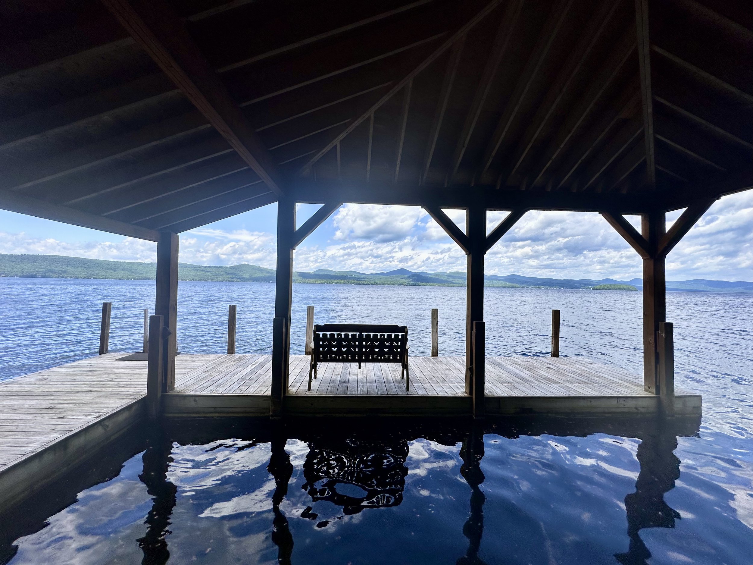 A wooden dock with a bench under a roof, overlooking a lake with mountains in the distance and a partly cloudy sky.