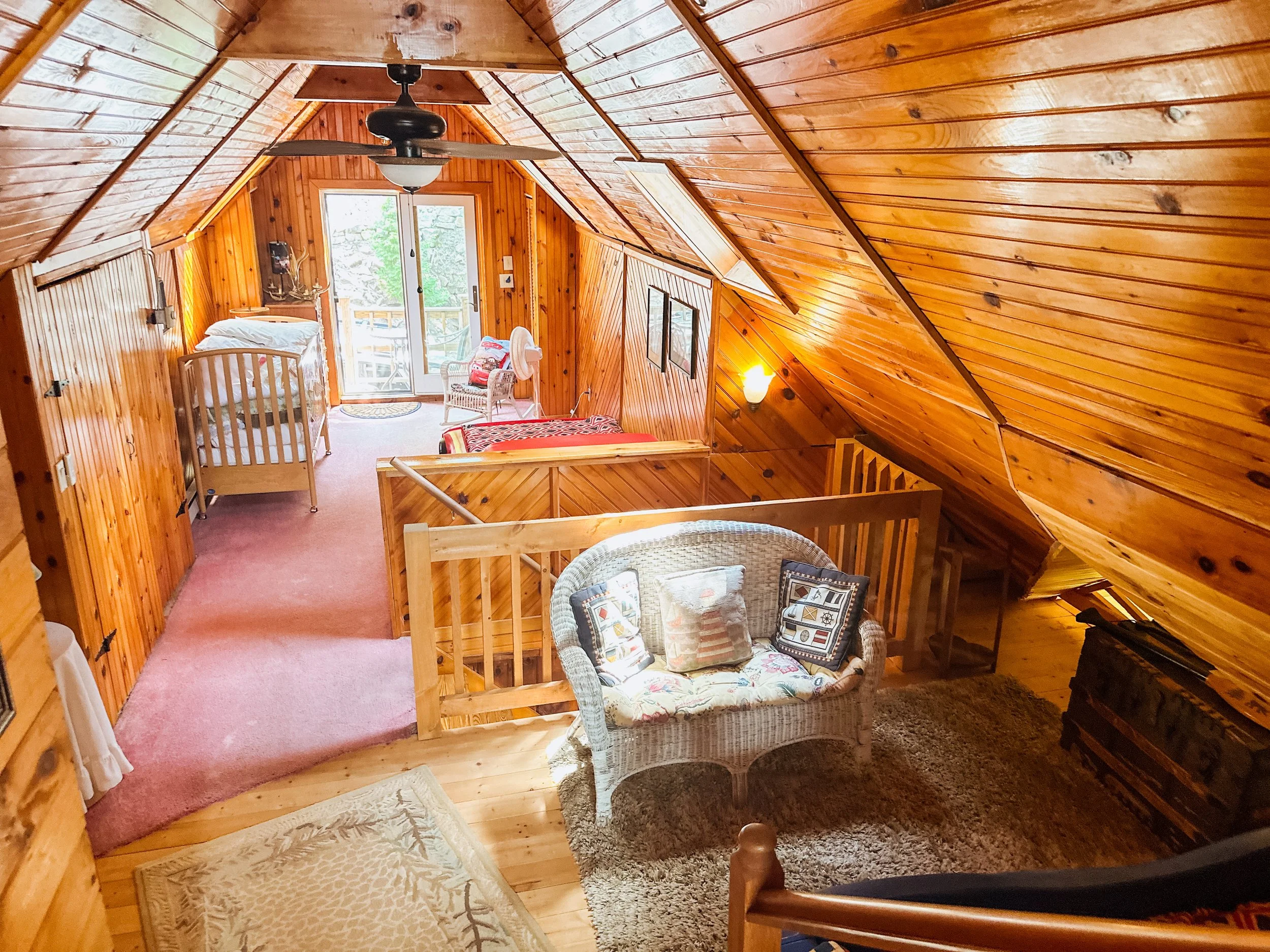Interior of a cozy wooden cabin bedroom with a sloped ceiling, a window showing trees outside, a crib, a wicker chair with pillows, and a small bench, warm lighting, and a ceiling fan.