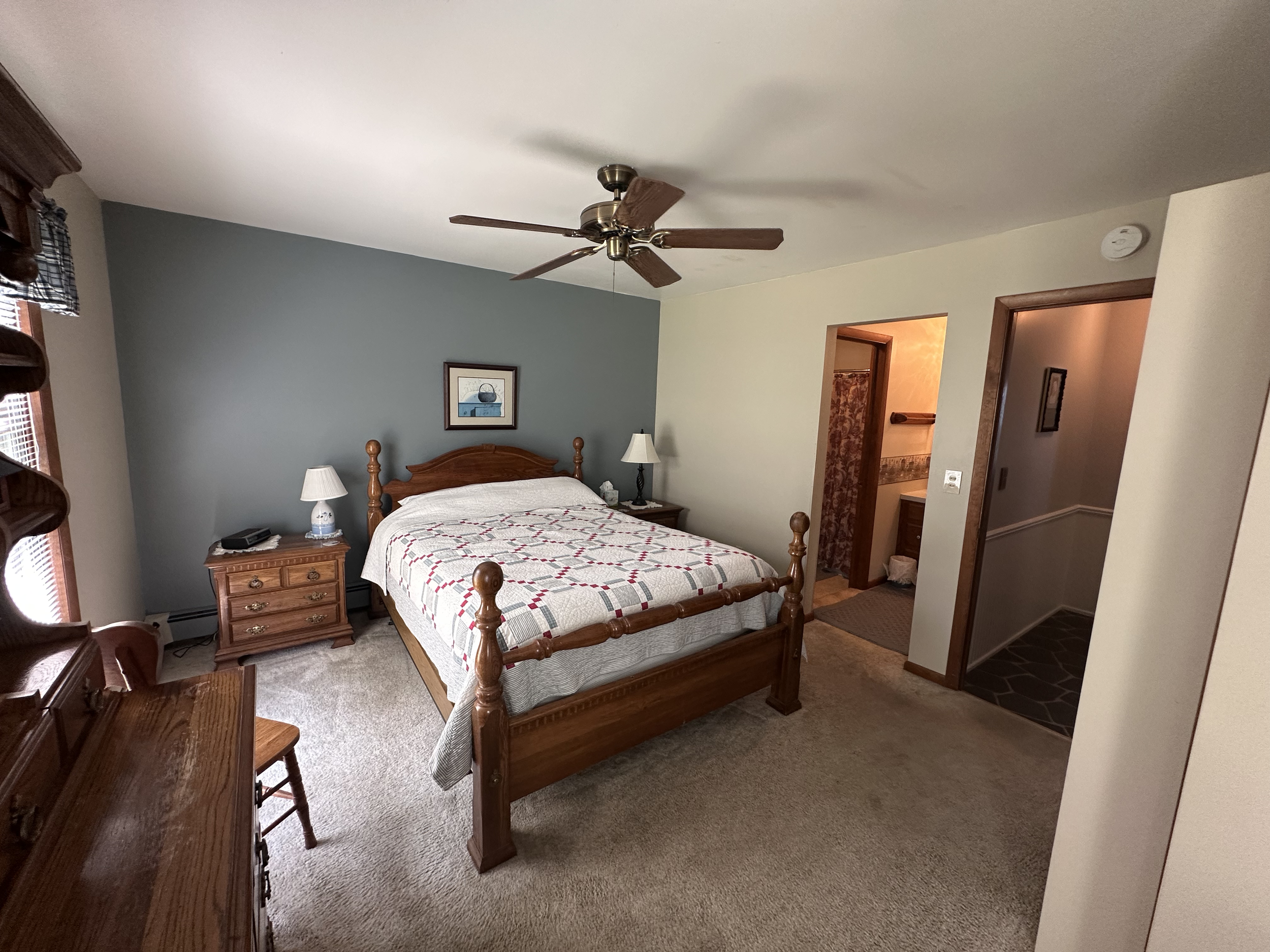 A bedroom with a wooden bed frame, a quilted bedspread, two nightstands with lamps, a ceiling fan, and an attached bathroom visible through an open doorway.