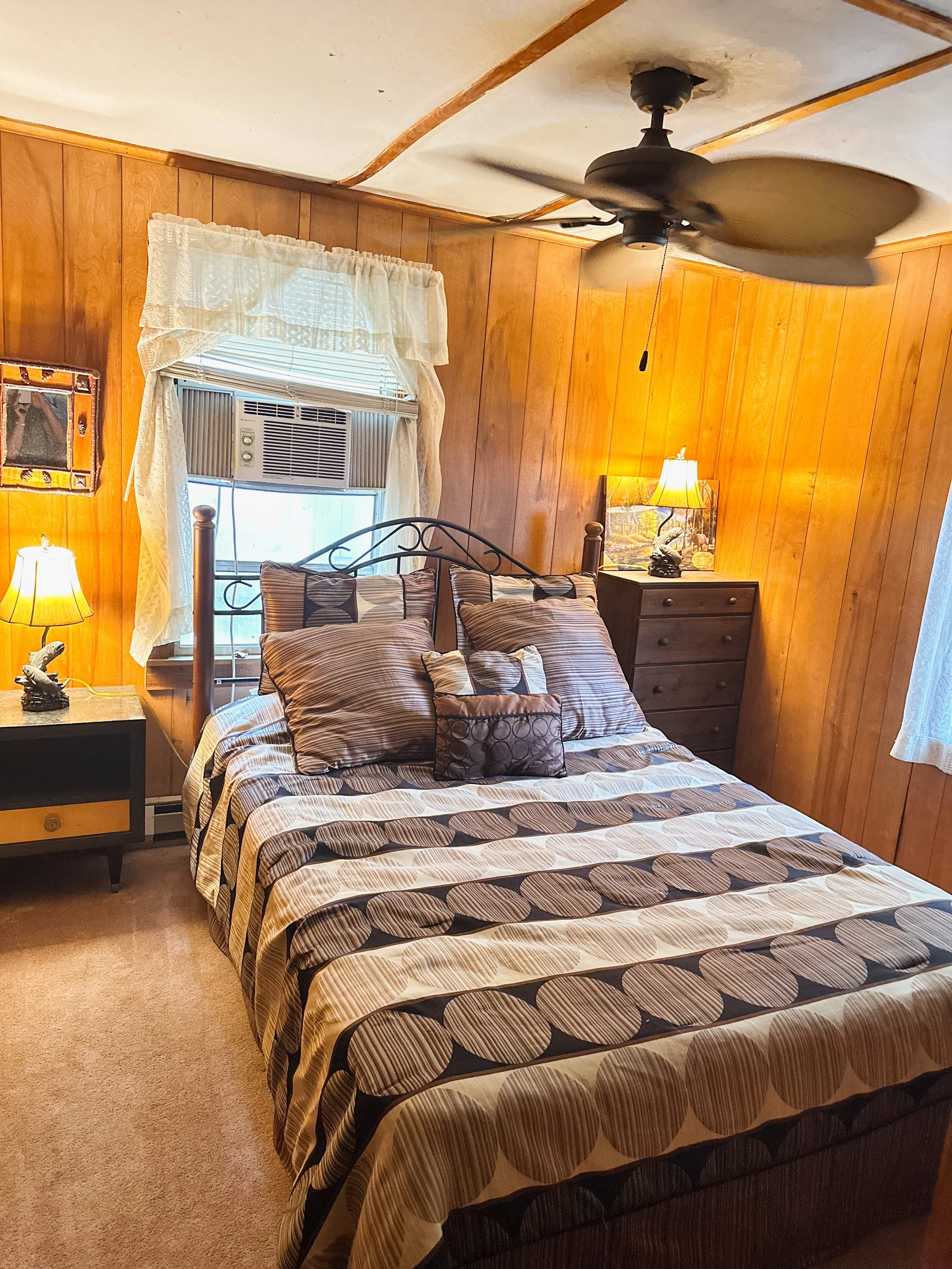 Bedroom with wooden paneled walls, a bed with a striped comforter and pillows, a nightstand with a lamp, a window with lace curtains and an air conditioning unit, ceiling fan, and a lamp on the nightstand.
