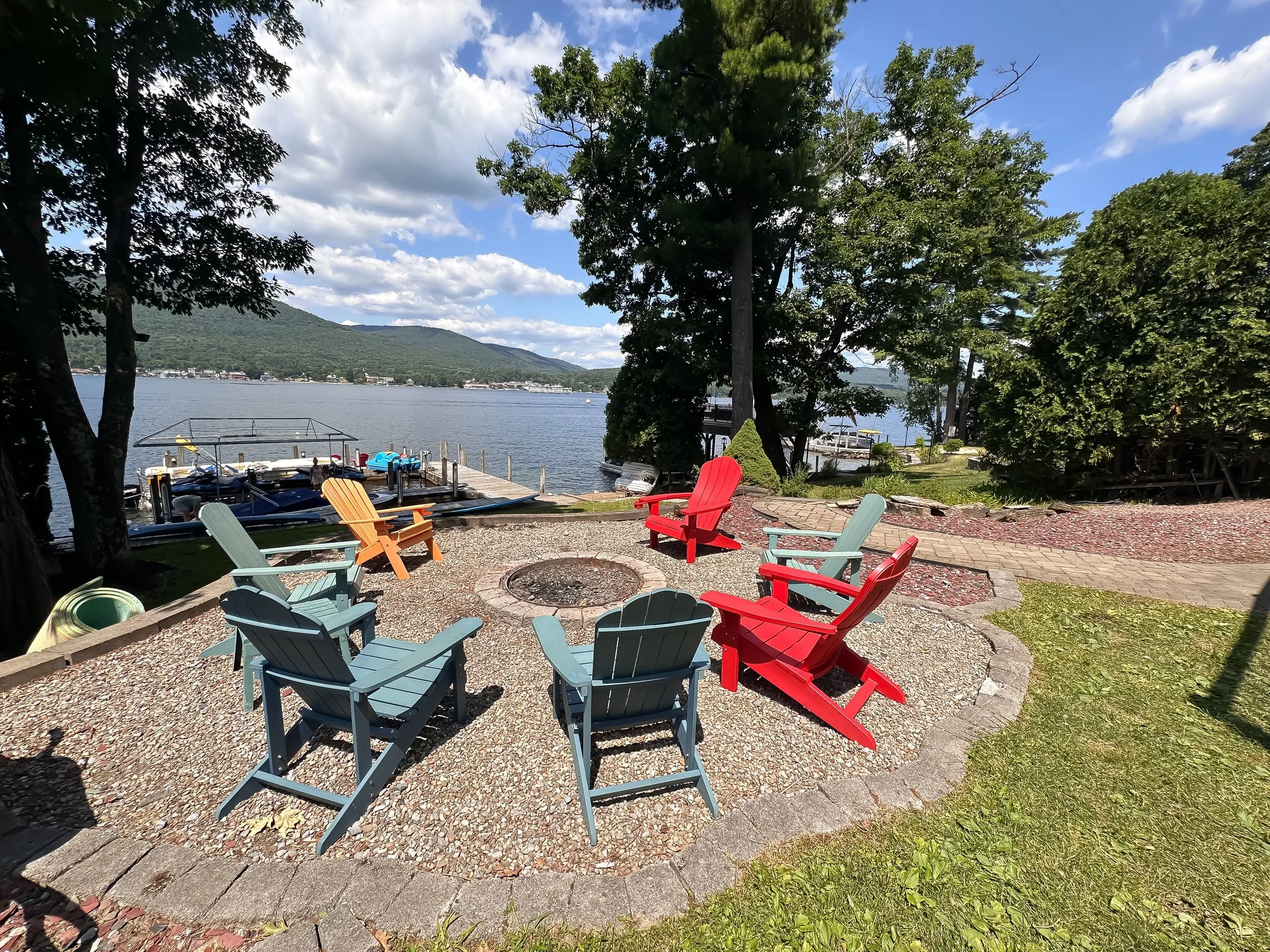 Colorful Adirondack chairs arranged around a fire pit on a gravel patio overlooking a lake with trees and mountains in the background.
