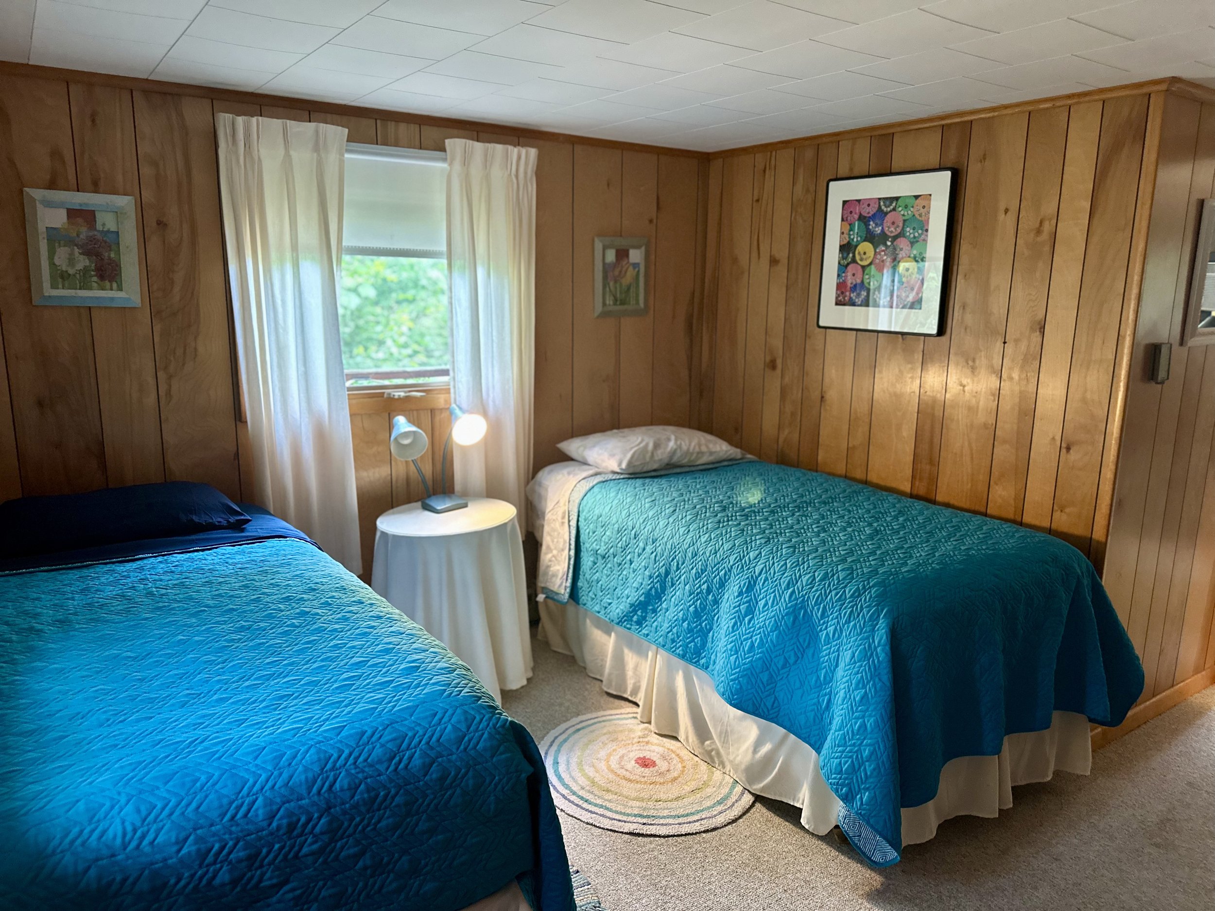 A cozy bedroom with two beds covered in blue quilts, wooden paneled walls, a small round table with a lamp, framed pictures on the wall, and a window with cream curtains overlooking greenery.