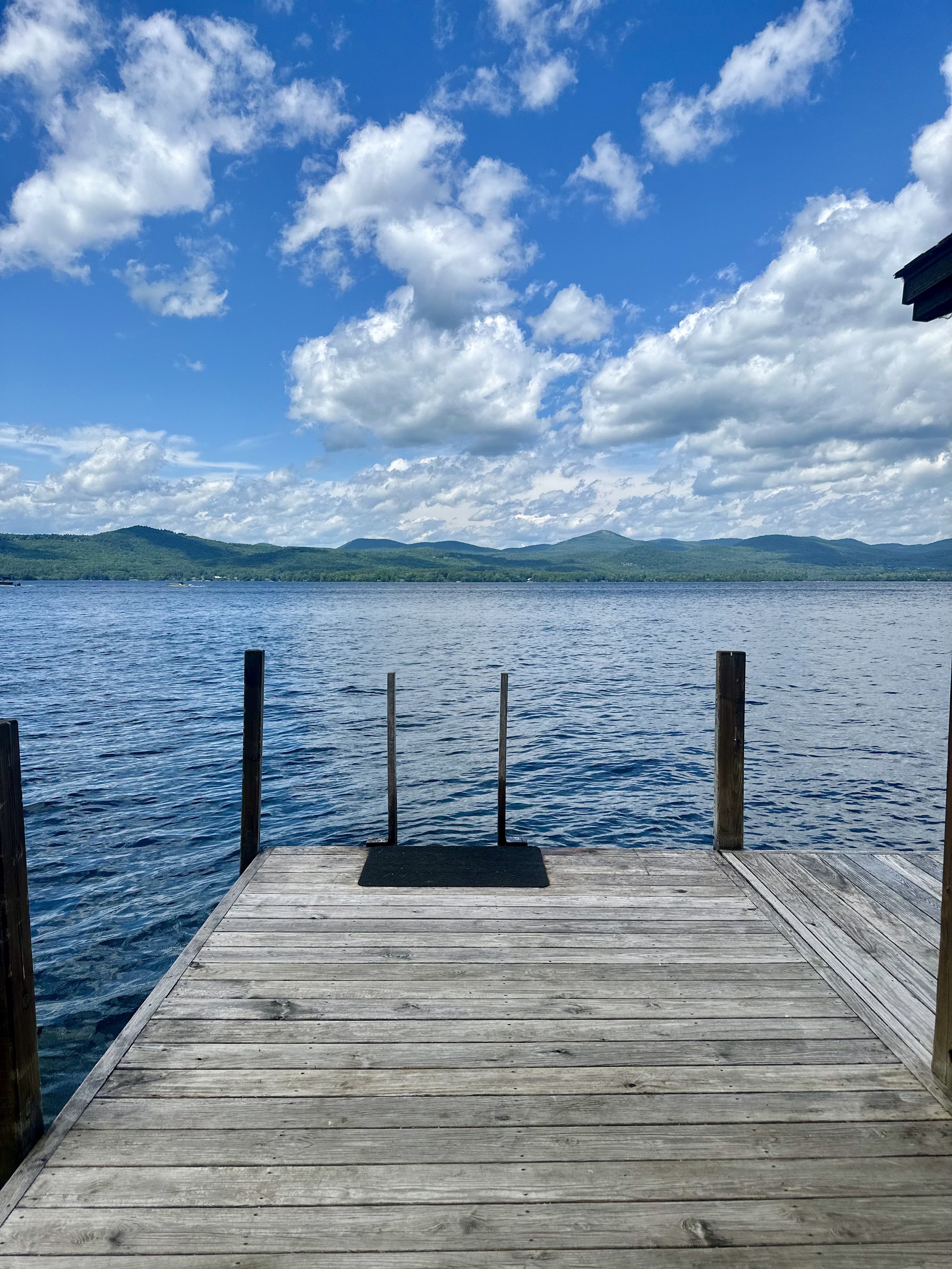 A wooden dock extending over a lake with mountains in the distance under a partly cloudy sky.
