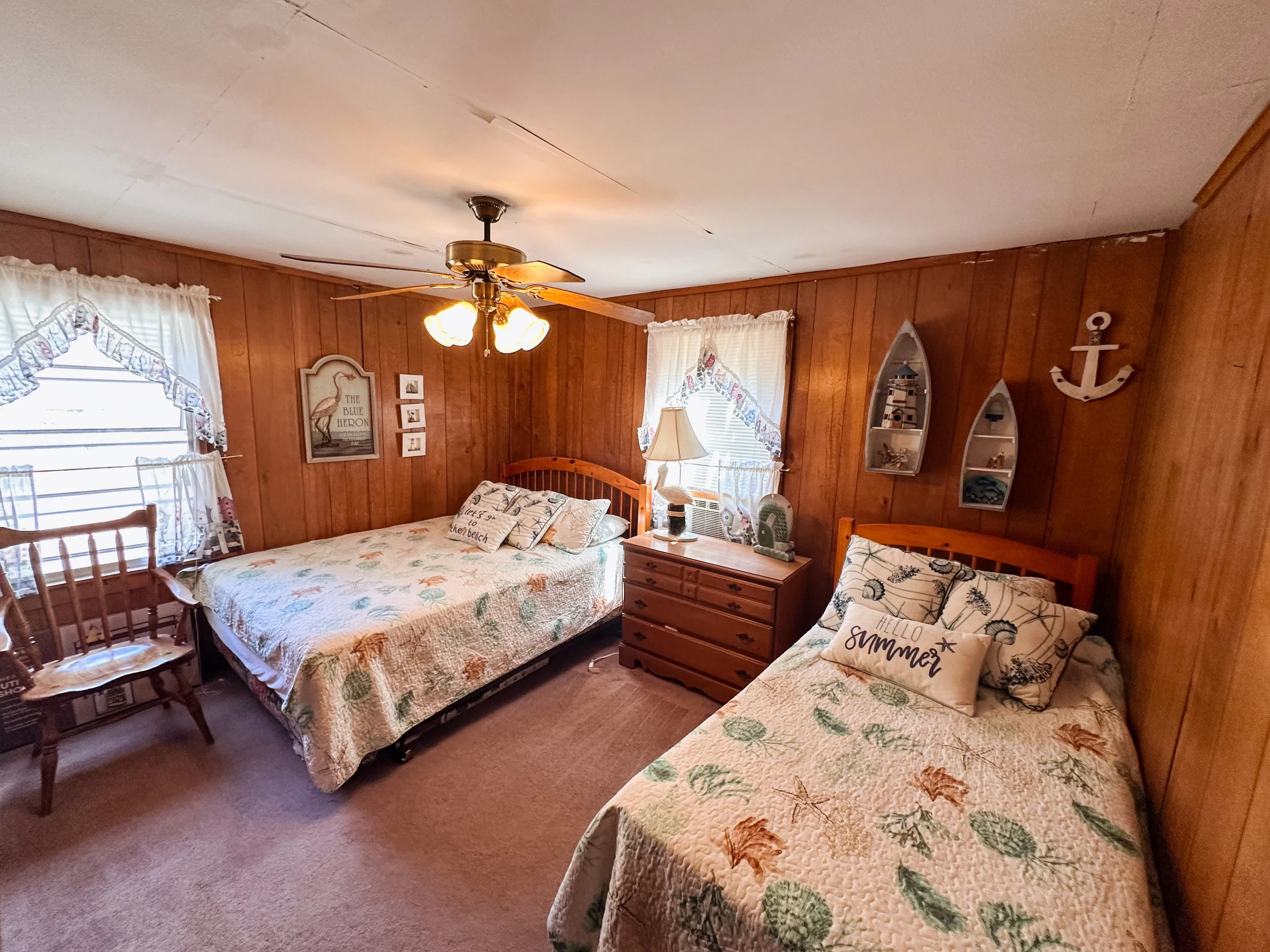 A cozy bedroom with wood-paneled walls, two beds with floral quilts, decorative pillows, and a ceiling fan with lights. There are windows with valances, a wooden dresser, a lamp, and nautical-themed wall decorations.