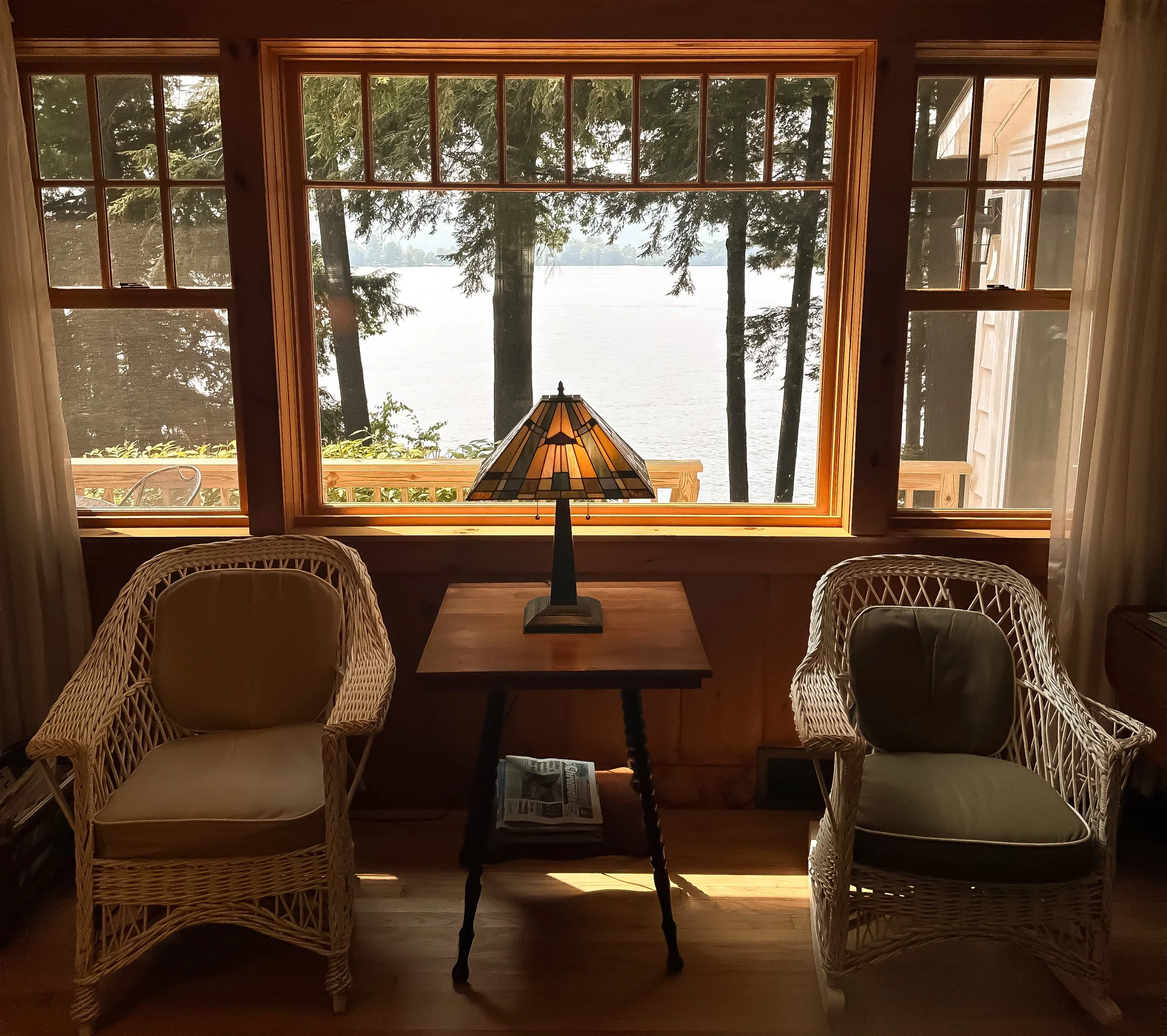 Interior of a cozy room with two wicker chairs and a wooden table with a stained glass lamp, overlooking a lake through large front windows with trees outside.