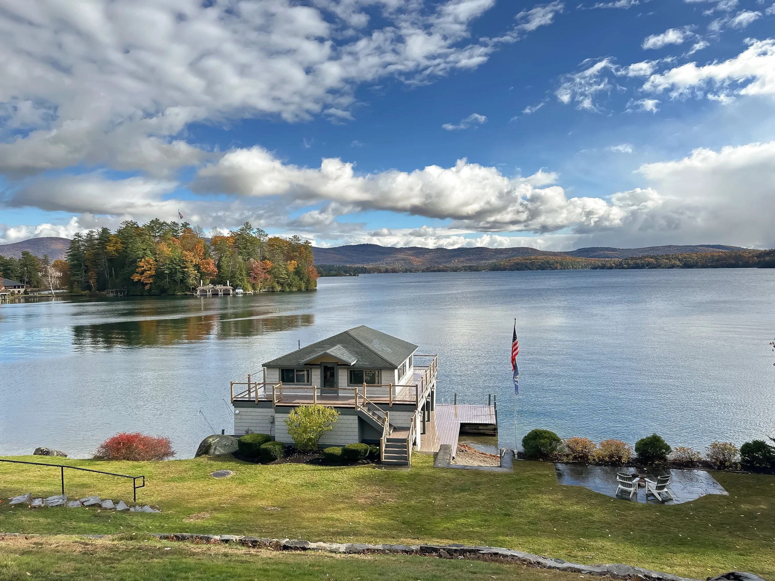 A lakeside house on a grassy yard with a deck and outdoor seating, overlooking a calm lake with a wooded island and hills in the distance, under a partly cloudy sky.