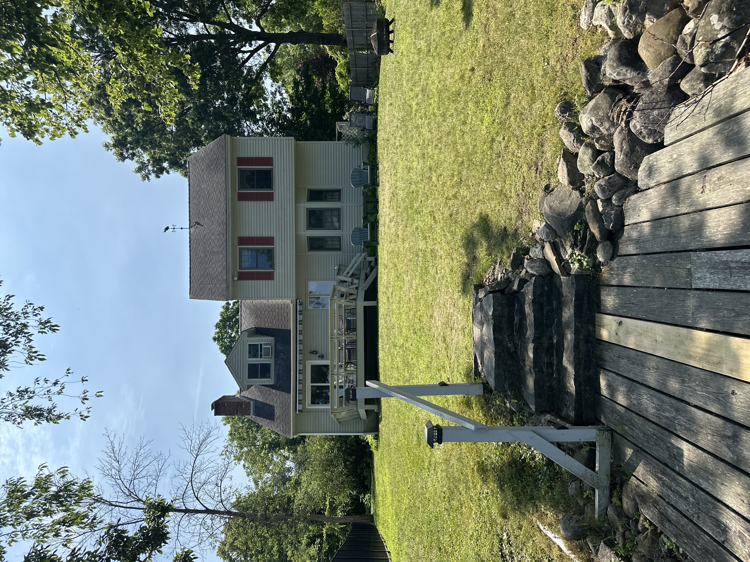 Backyard with a wooden deck, stone border, green grass, trees, and a two-story cream-colored house with red window shutters.