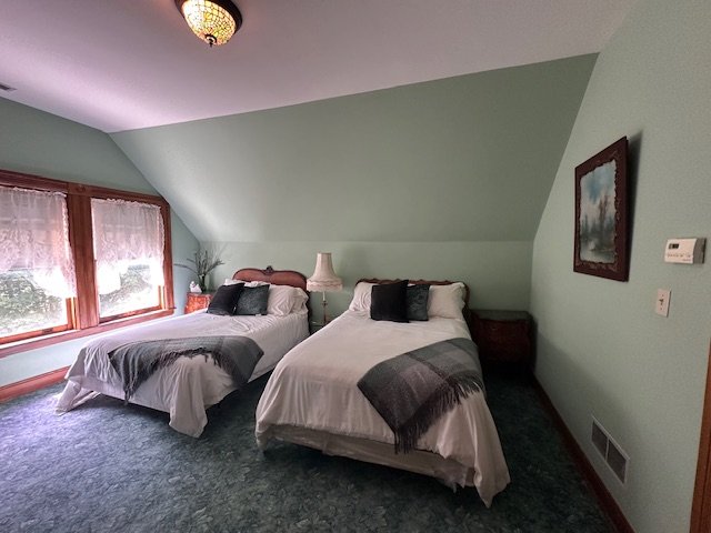 Two beds in a room with green walls, each with pillows and blankets, a window with lace curtains, a nightstand with a lamp, and a framed picture on the wall.