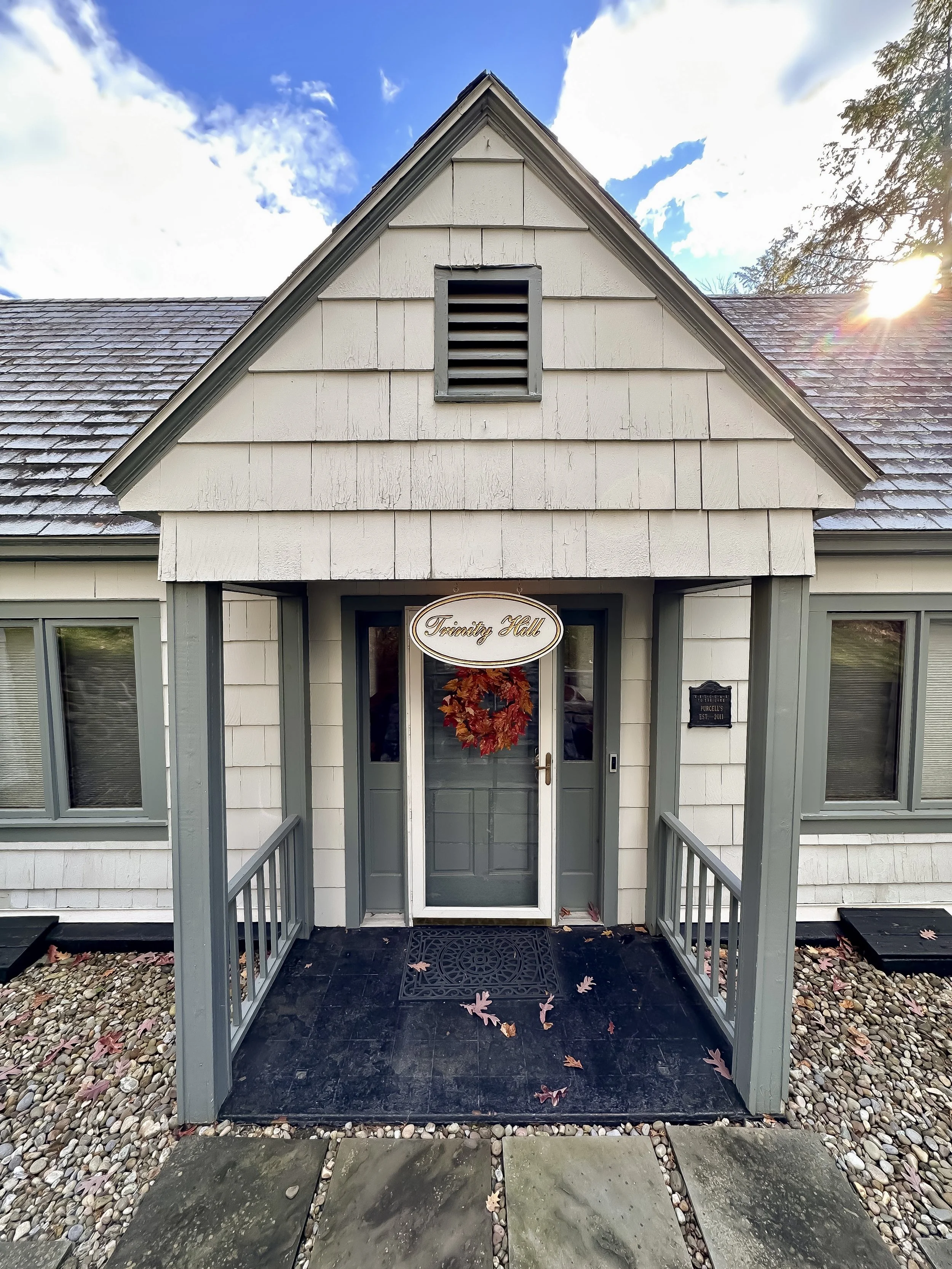 Front porch of a house with a white door decorated with a fall wreath; sign above the door reads "Tranquil Hollow"; small black plaque to the right of the door; gray trim and railings; pebble and stone pathway; autumn leaves on the porch and pathway;