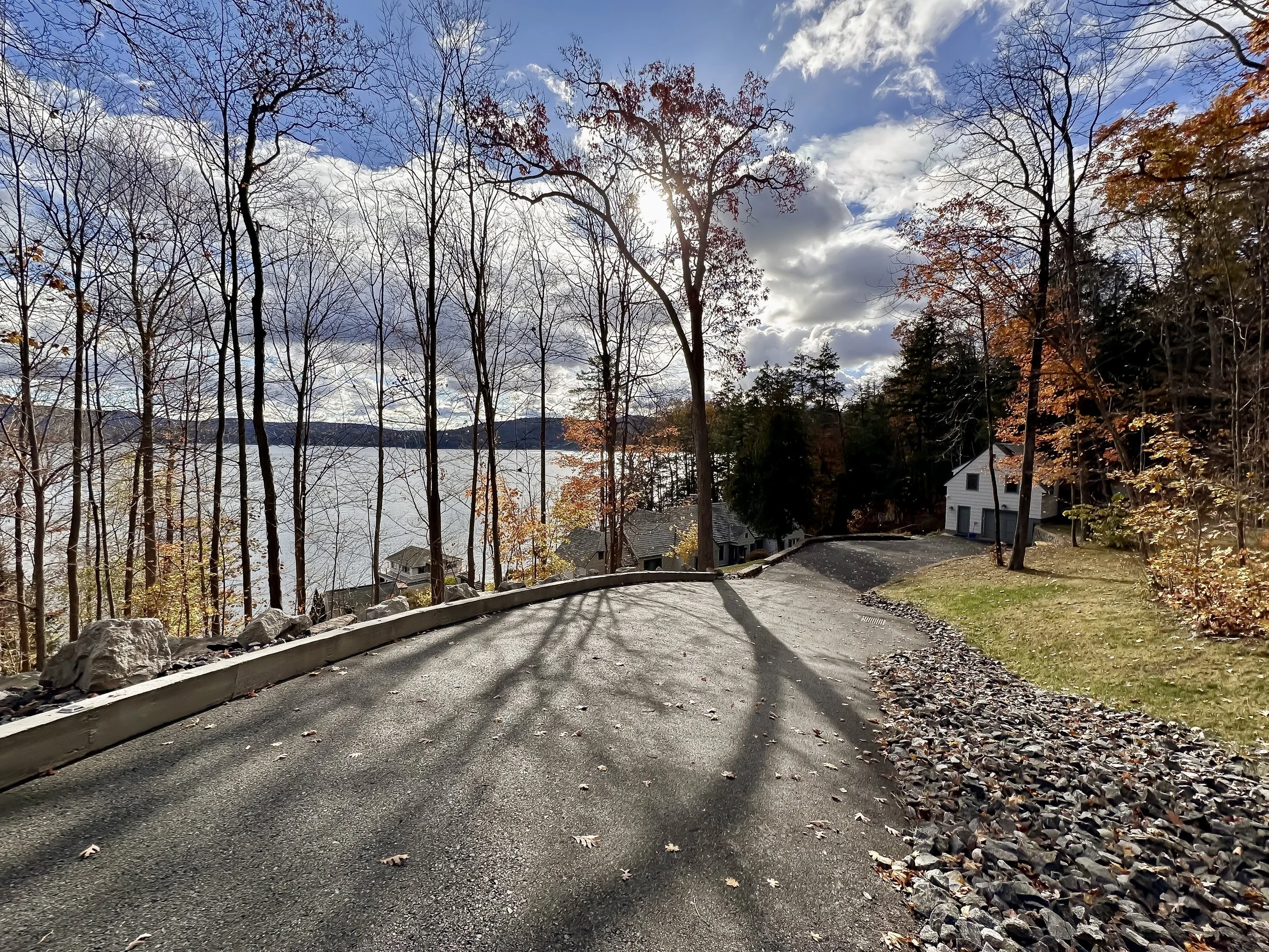 A winding road in autumn with leafless and partially leafed trees on either side, leading to a house near a lake, with cloudy sky overhead.
