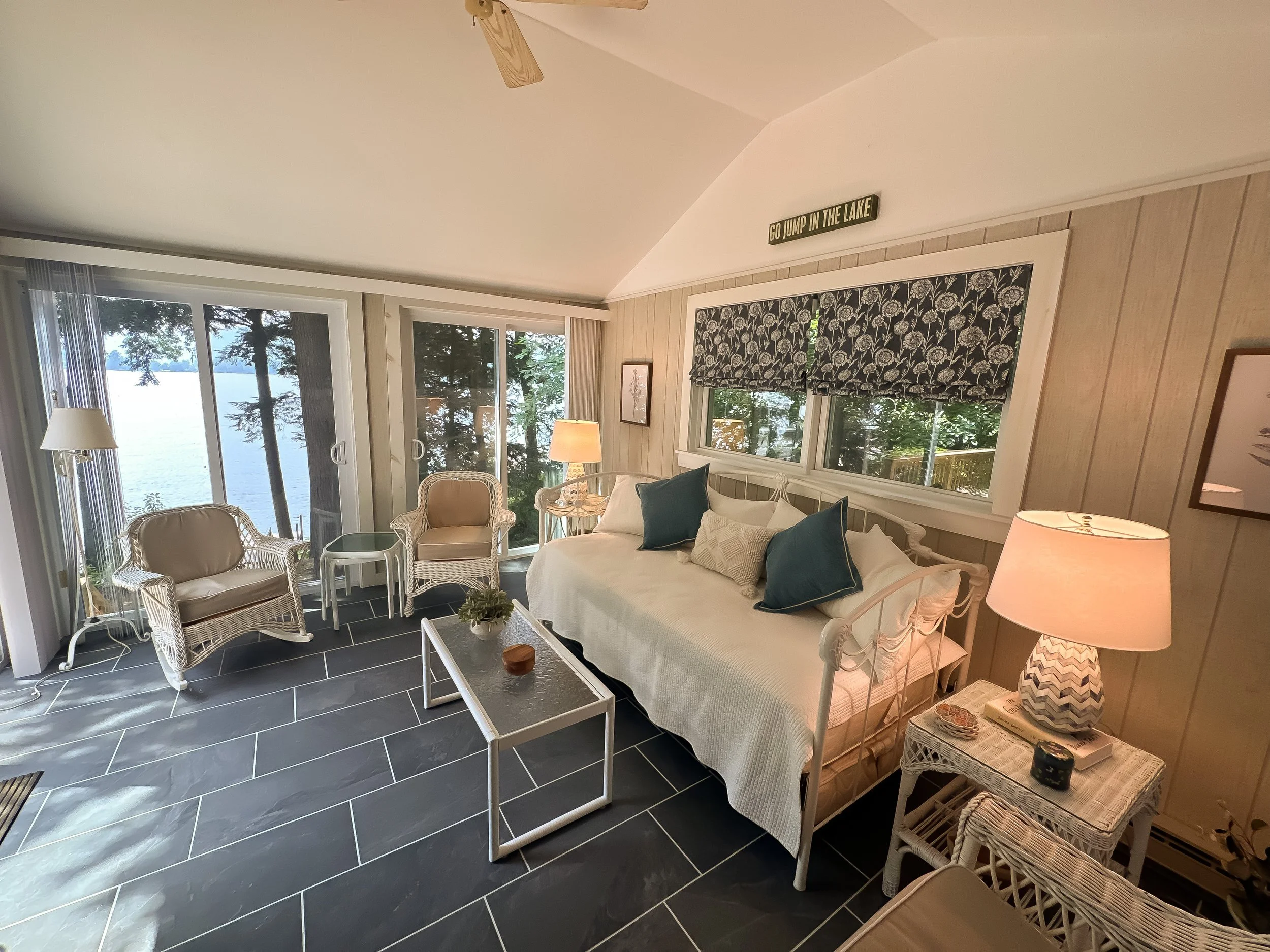 Living room with large glass windows overlooking a lake, white furniture including a daybed with teal and white pillows, two wicker chairs, a white coffee table, and side tables with lamps.