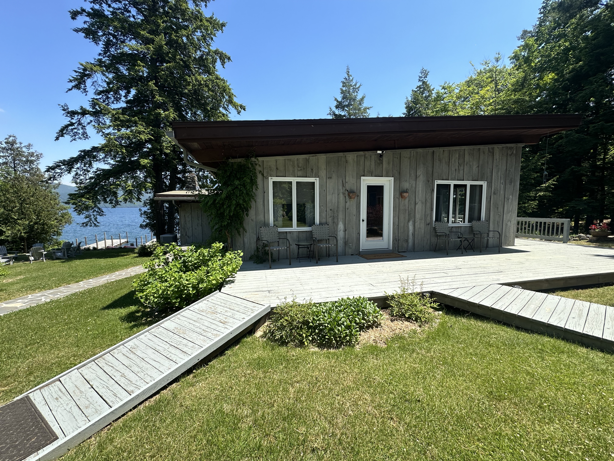 Wooden house by a lake with a deck, surrounded by green grass, trees, and blue sky.