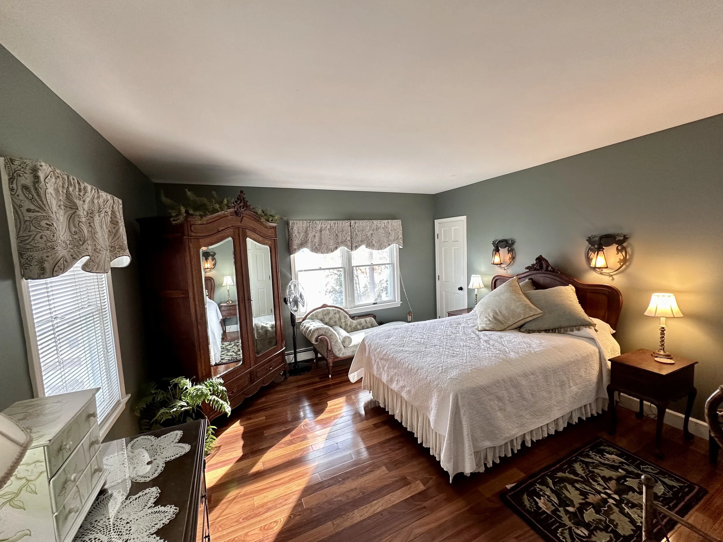 A cozy bedroom with a large bed, white quilt, and decorative pillows, wooden furniture including a wardrobe, nightstands with lamps, a vintage settee, and a potted plant, with windows allowing natural light to fill the room.