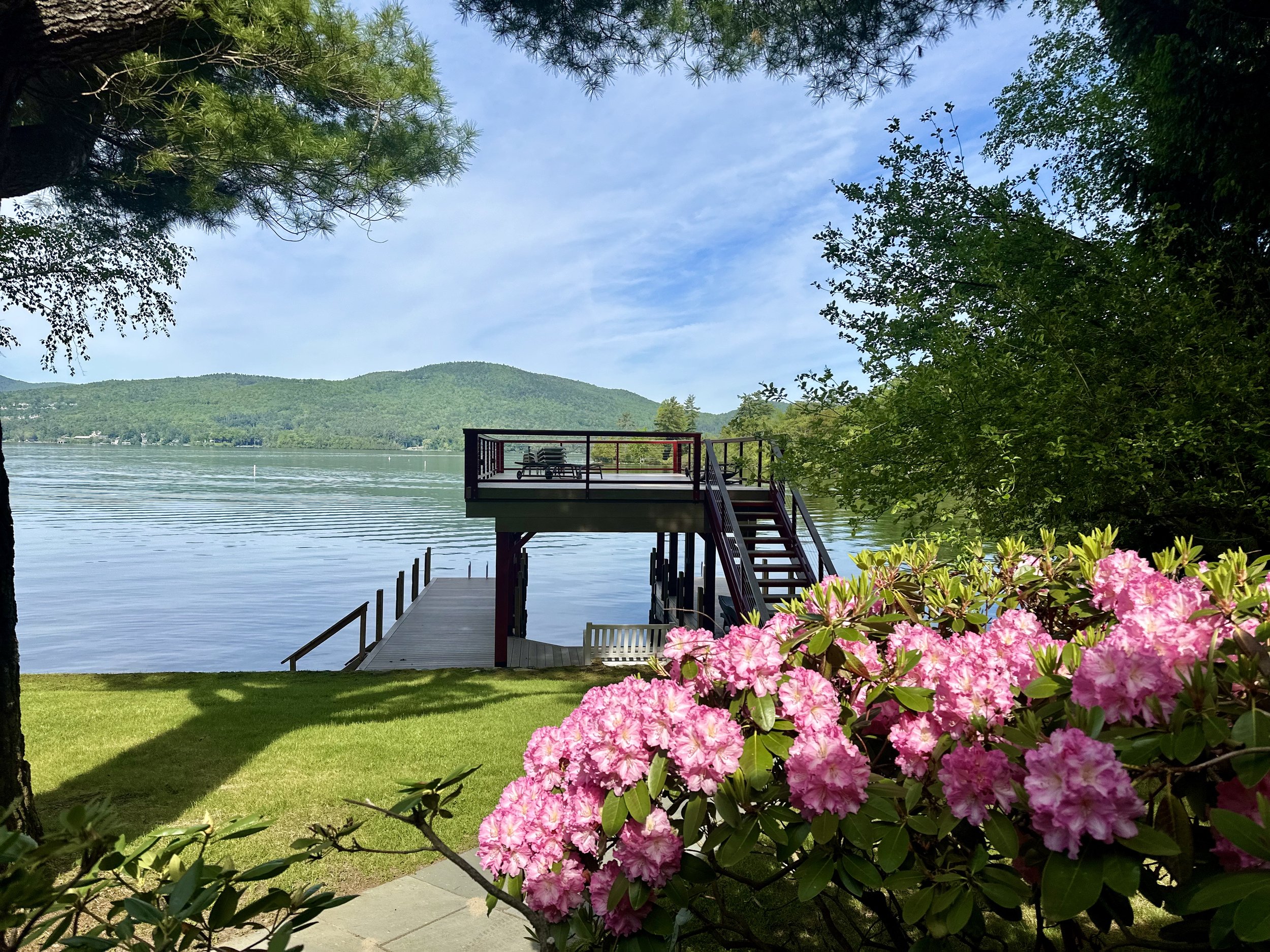 A lakeside scene featuring a wooden deck with stairs leading down to the water, pink rhododendron flowers in the foreground, green grass, trees, calm lake water, distant green hills, and a partly cloudy blue sky.
