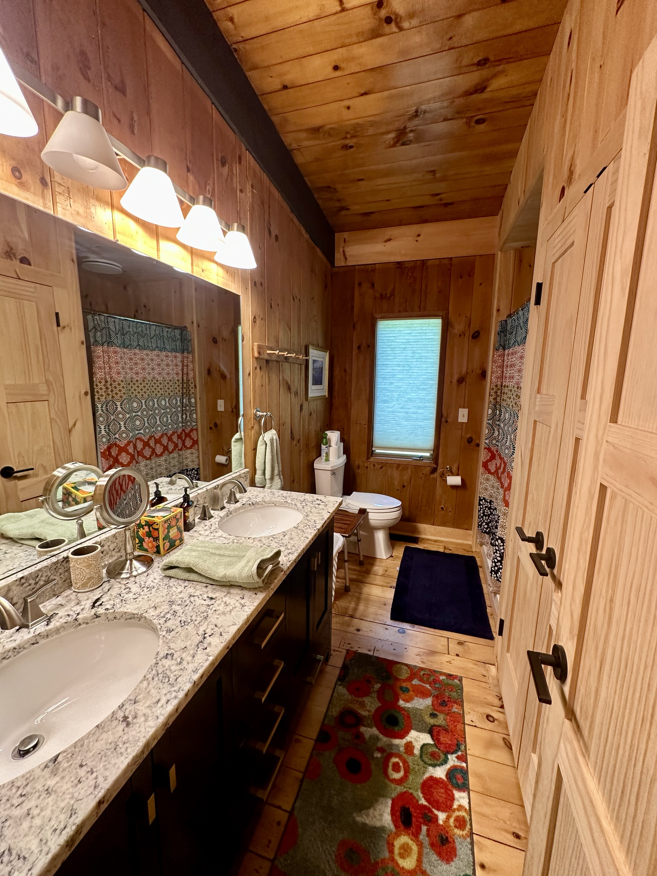 Bathroom with wooden walls and ceiling, granite countertop with two sinks, colorful patterned rugs, mirror, decorative items, toilet, and window.