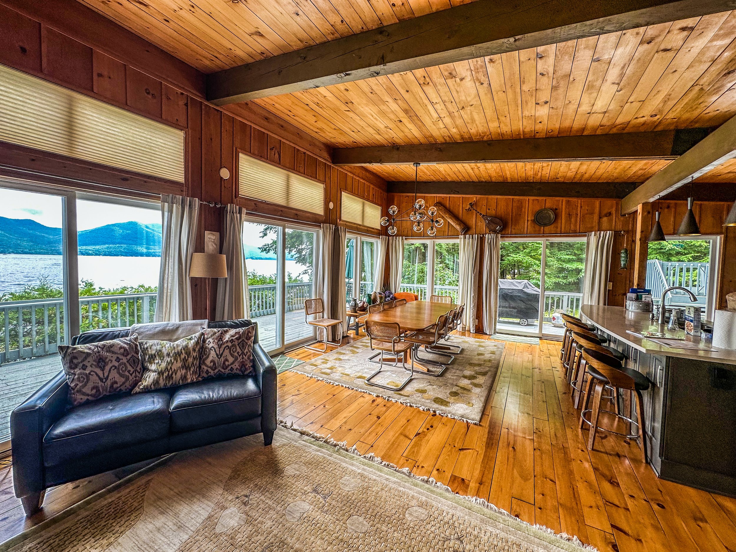 Interior of a wooden cabin living room with large windows overlooking a lake, featuring a black sofa with patterned pillows, a dining table with chairs, a kitchen island with bar stools, and a deck outside.