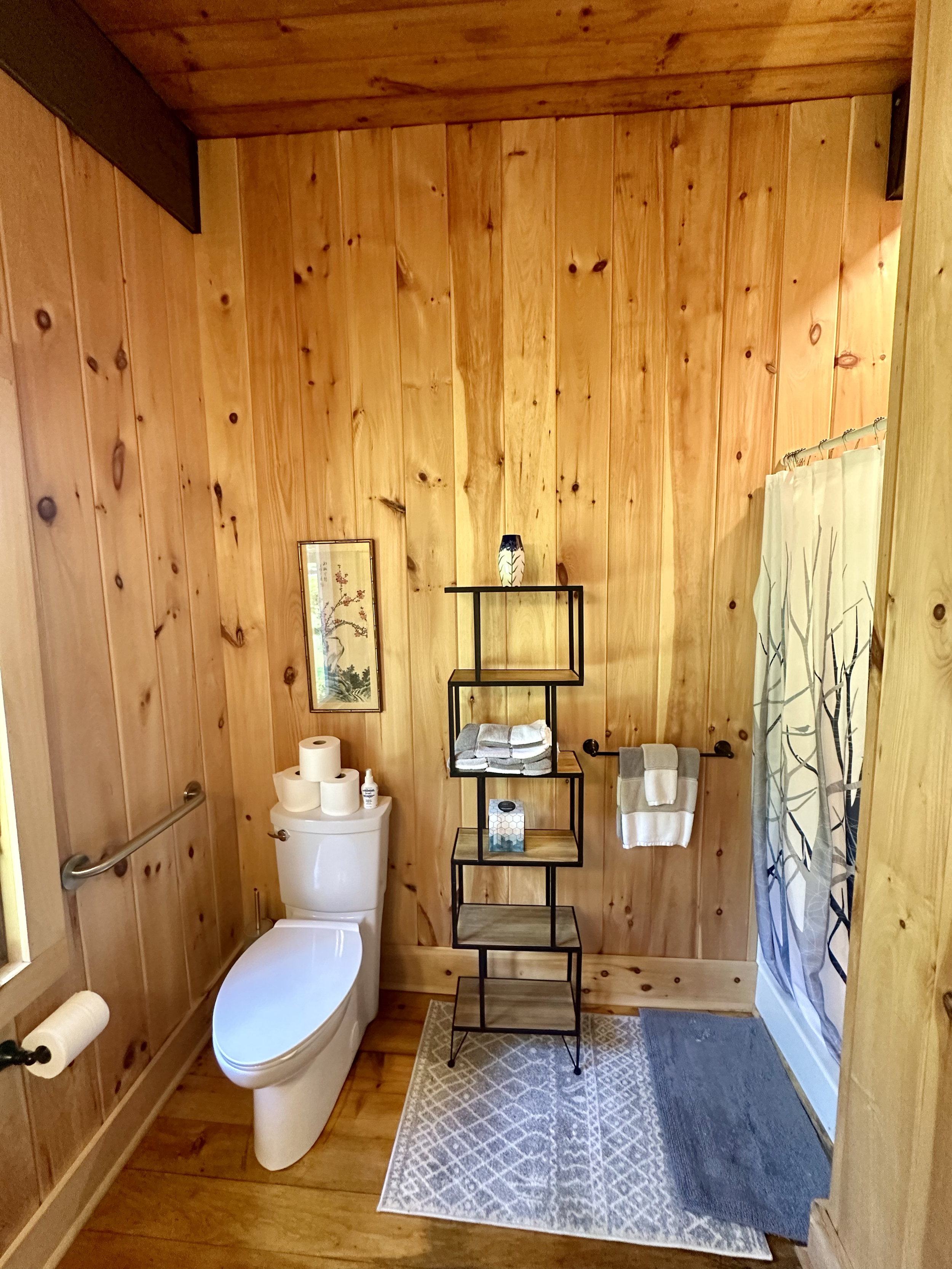 A bathroom with wooden walls and ceiling, a white toilet with toilet paper rolls and a small bottle on top, a black metal shelving unit with folded towels and decorative items, a towel hanging on a bar, a shower curtain with a tree branch design, a s