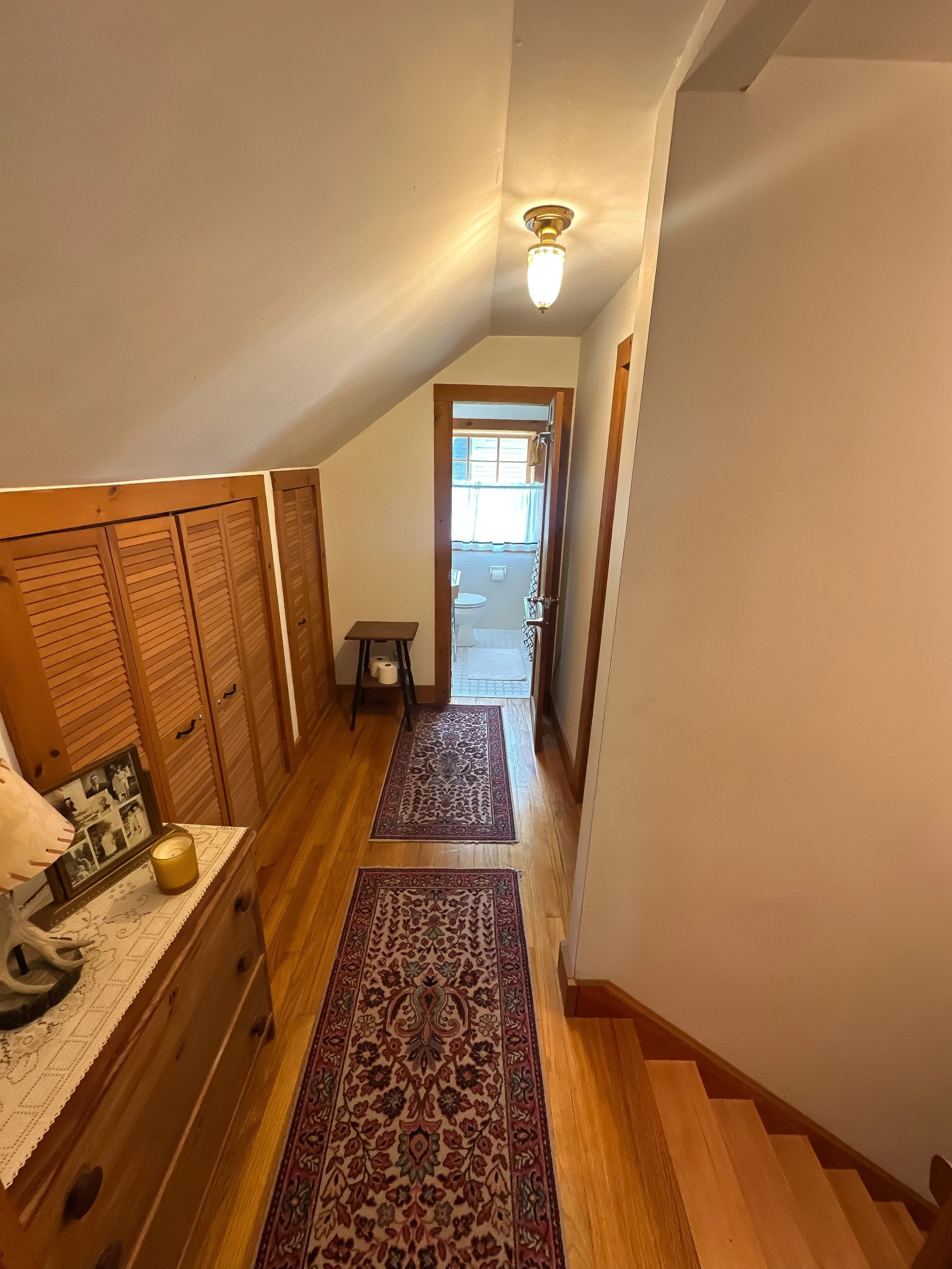View of a narrow wooden-floored hallway with rugs, a small table, and a bathroom with a window at the end.