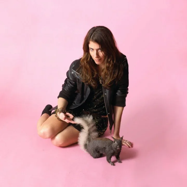 A woman sitting on a pink floor with her legs folded, wearing a black leather jacket and dress, holding the tail of a small, gray puppy, with a gray kitten nearby on the floor.