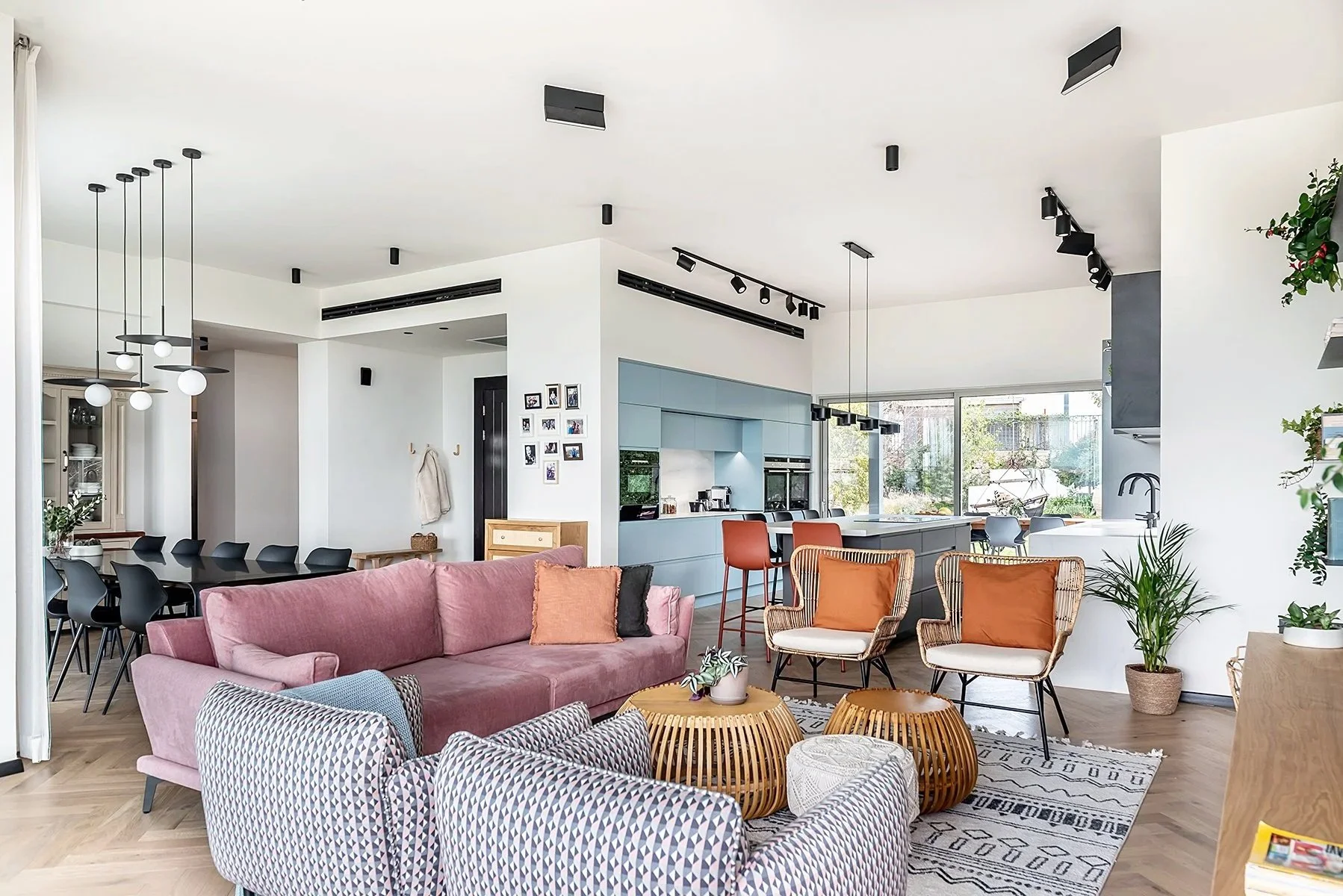 Open-concept living room and kitchen with modern furniture, colorful seating, large windows, plants, and light wood flooring.