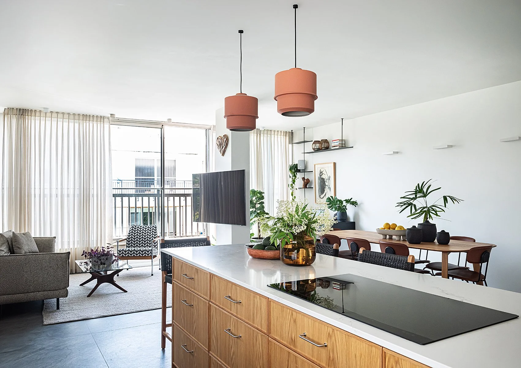 Modern open-concept living and dining area with a kitchen island, greenery, wooden furniture, and ample natural light from large windows with curtains.