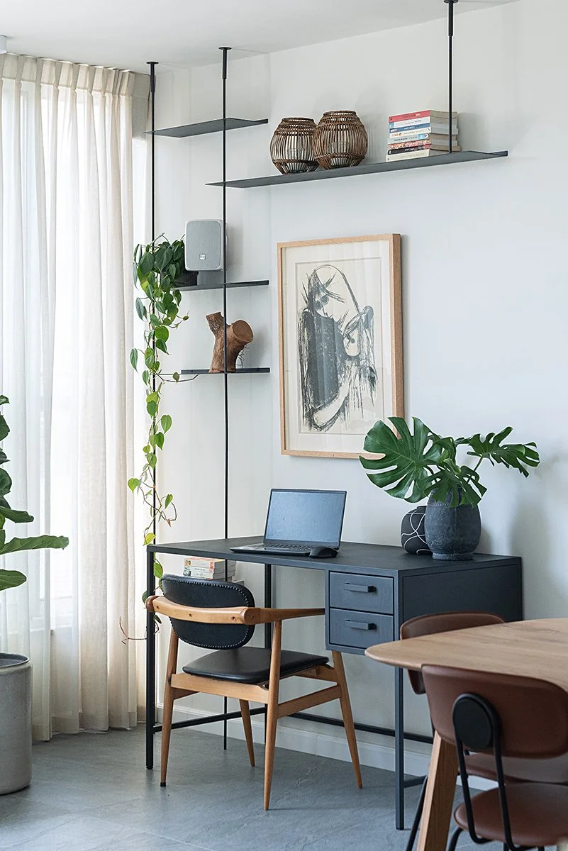Modern home office with black desk, wooden chair, laptop, green plants, framed artwork, and floating shelves with books and decorative baskets, next to a window with sheer curtains.