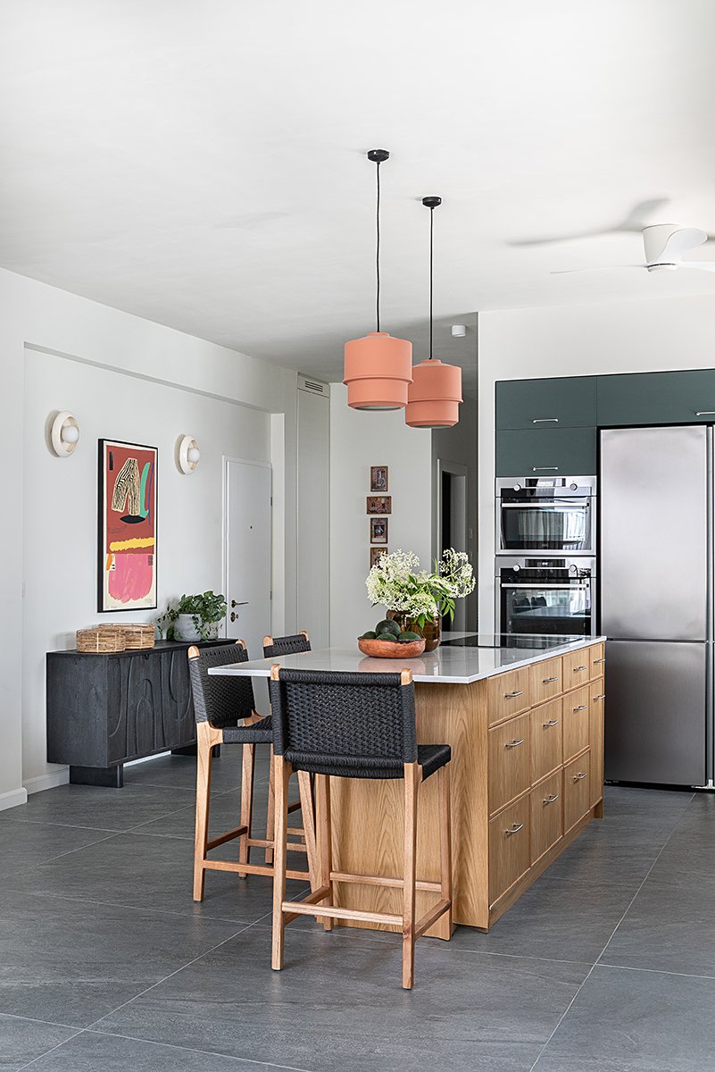 Modern kitchen with wooden island, black stools, stainless steel appliances, dark green cabinets, white walls, decorative wall art, pink pendant lights, and a white ceiling fan.
