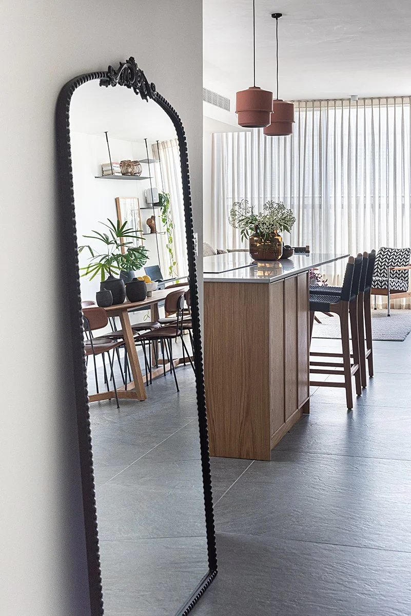 A tall, ornate mirror reflects a modern interior with a dining area and a kitchen island, decorated with plants, chairs, and pendant lights.