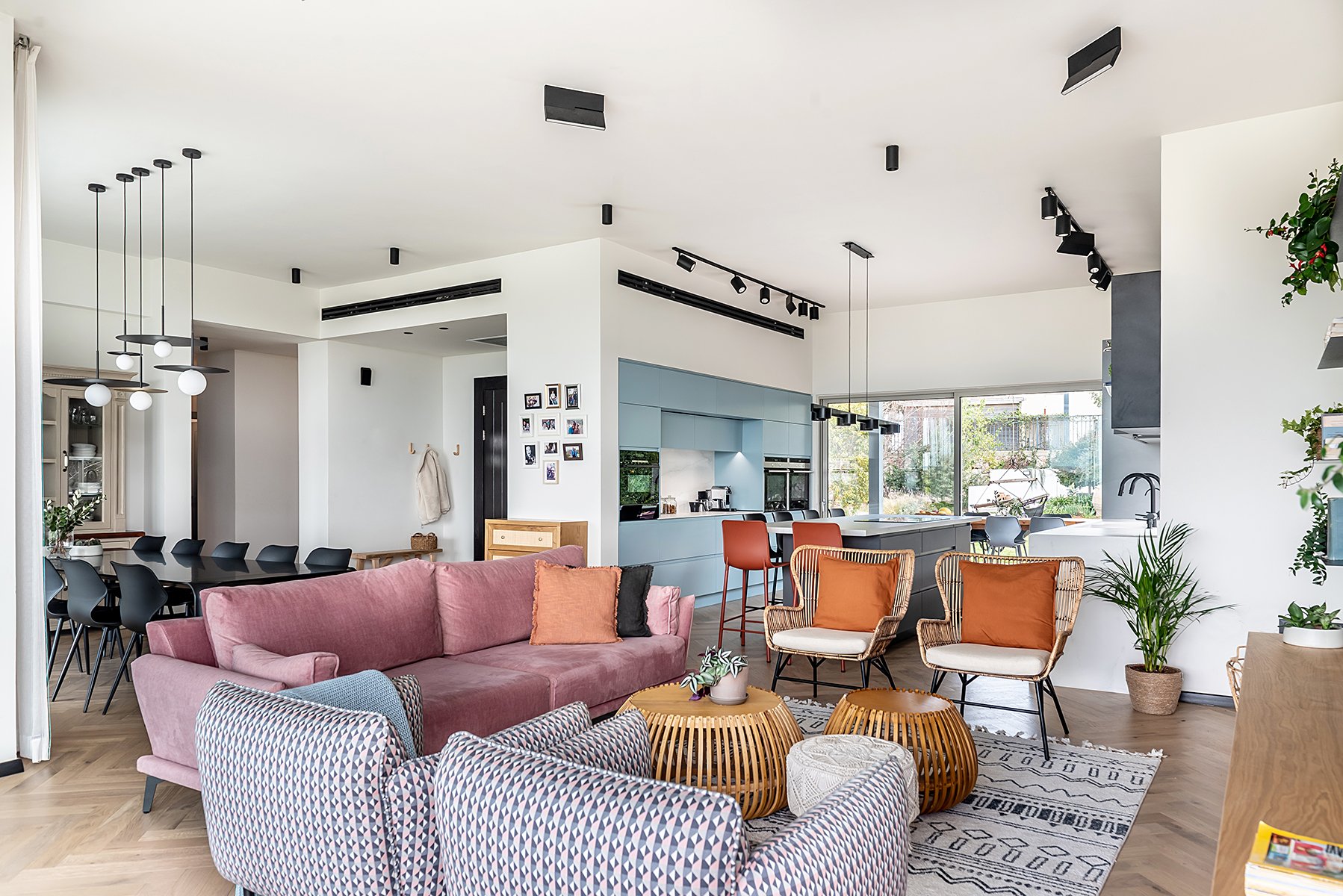 Open-concept living room and kitchen with modern furniture, colorful seating, large windows, plants, and light wood flooring.